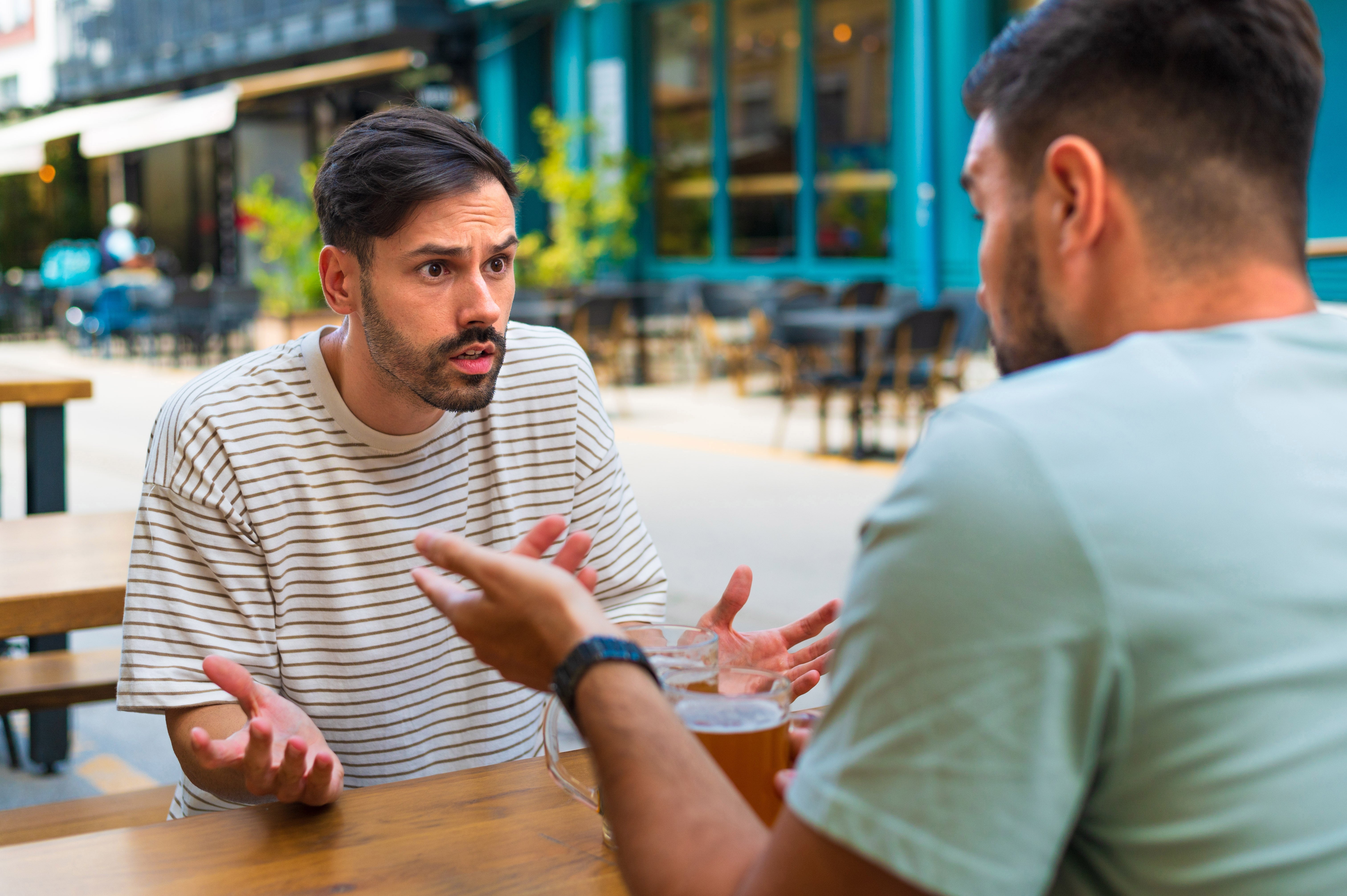 Two men at an outdoor cafe, engaged in an animated conversation. One is gesturing with his hands, conveying surprise or confusion