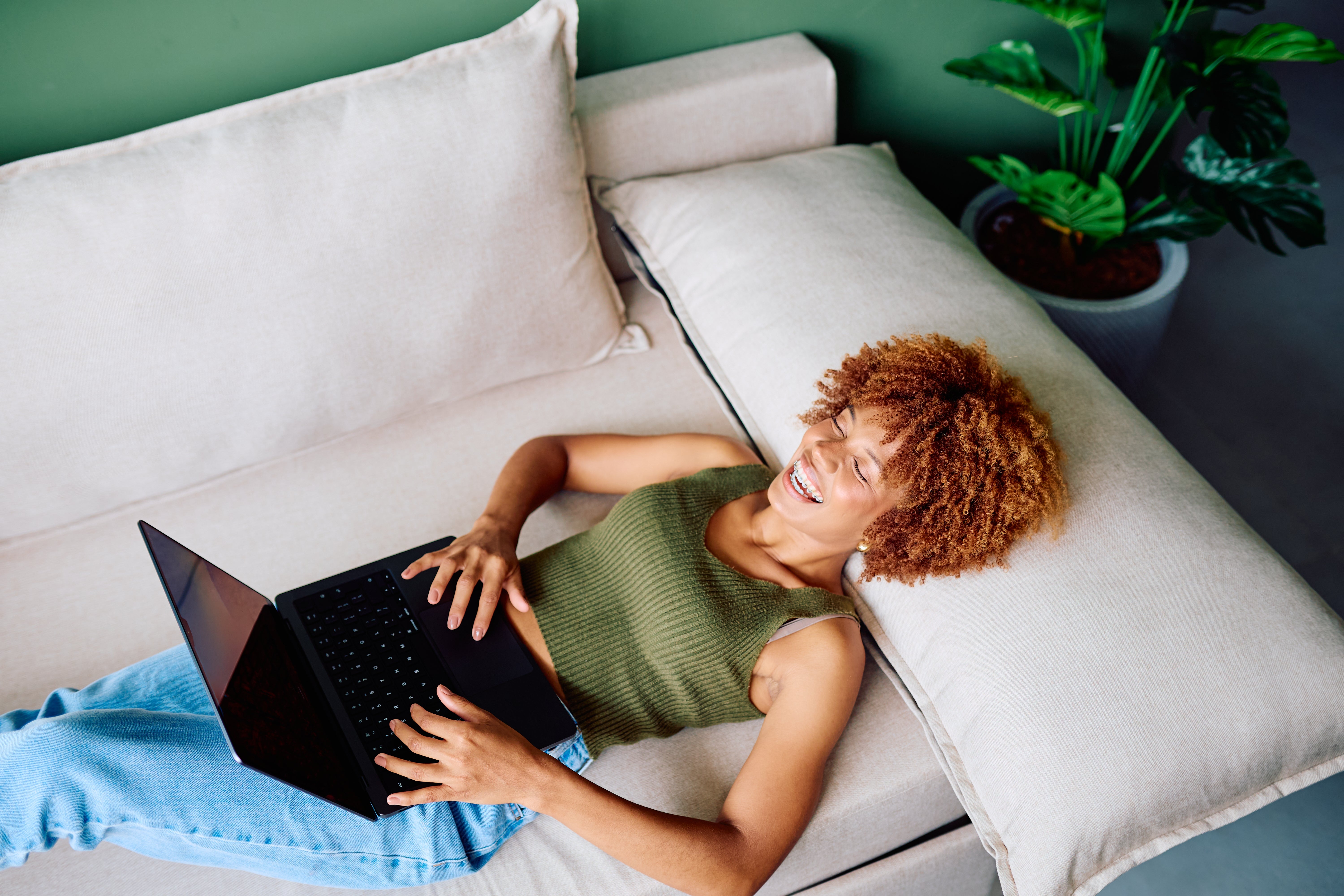 Person relaxing on a couch with a laptop, smiling and wearing a sleeveless top and jeans; a potted plant in the background