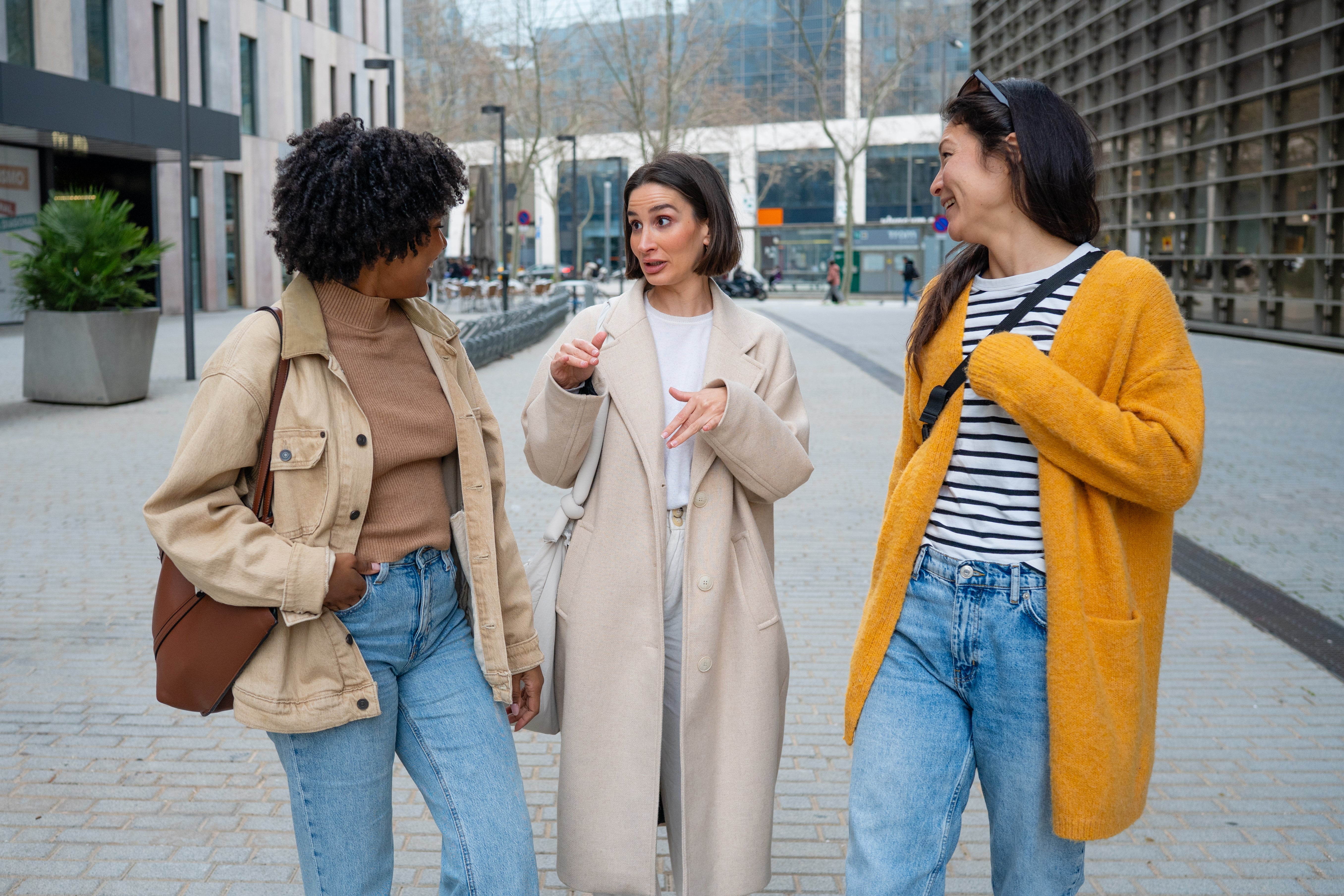 Three women walking and talking in a city street, dressed in casual spring outfits with denim jeans and lightweight jackets