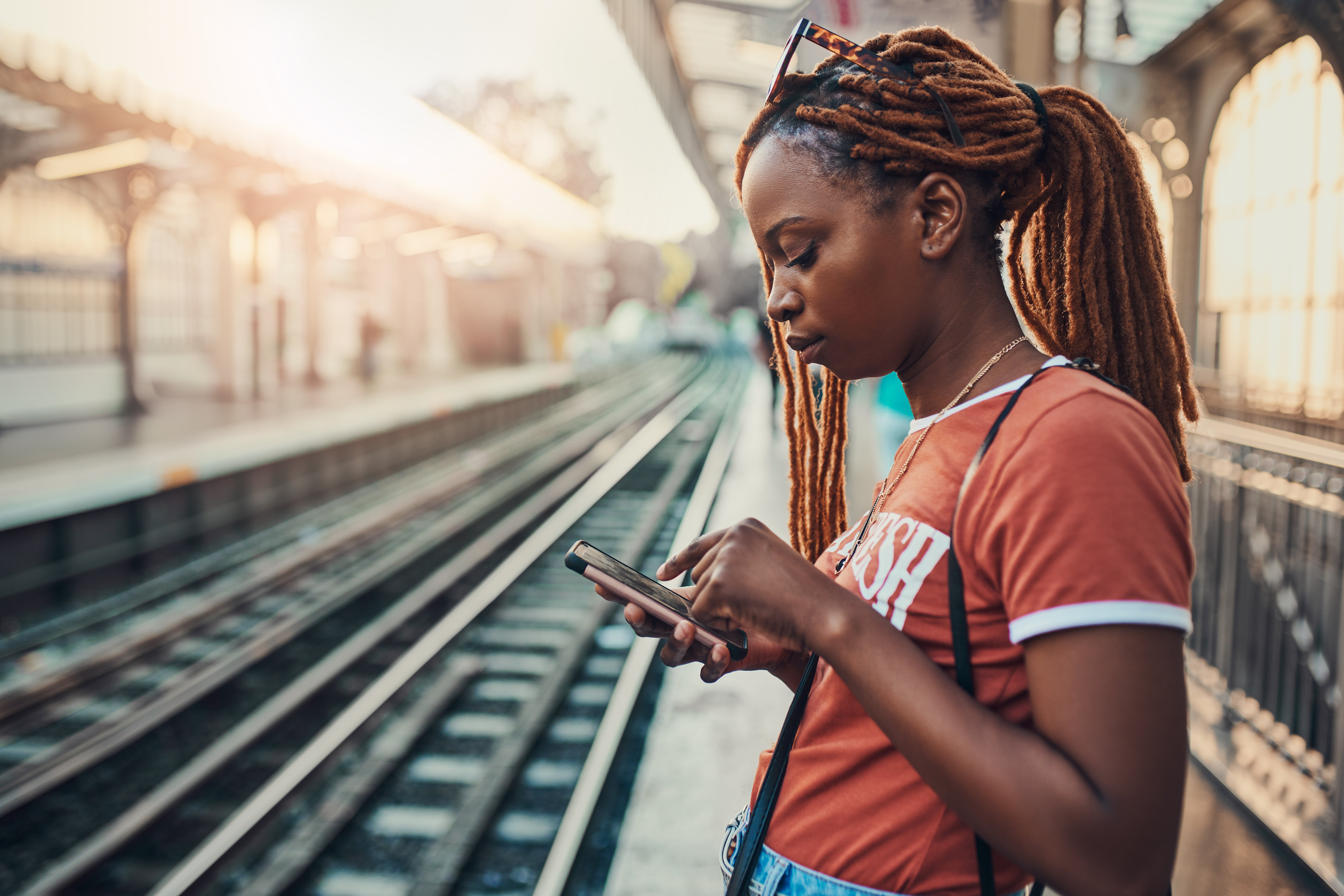 Person with braided hair standing at a train station platform, focused on their smartphone. Sunlight filters through the station roof