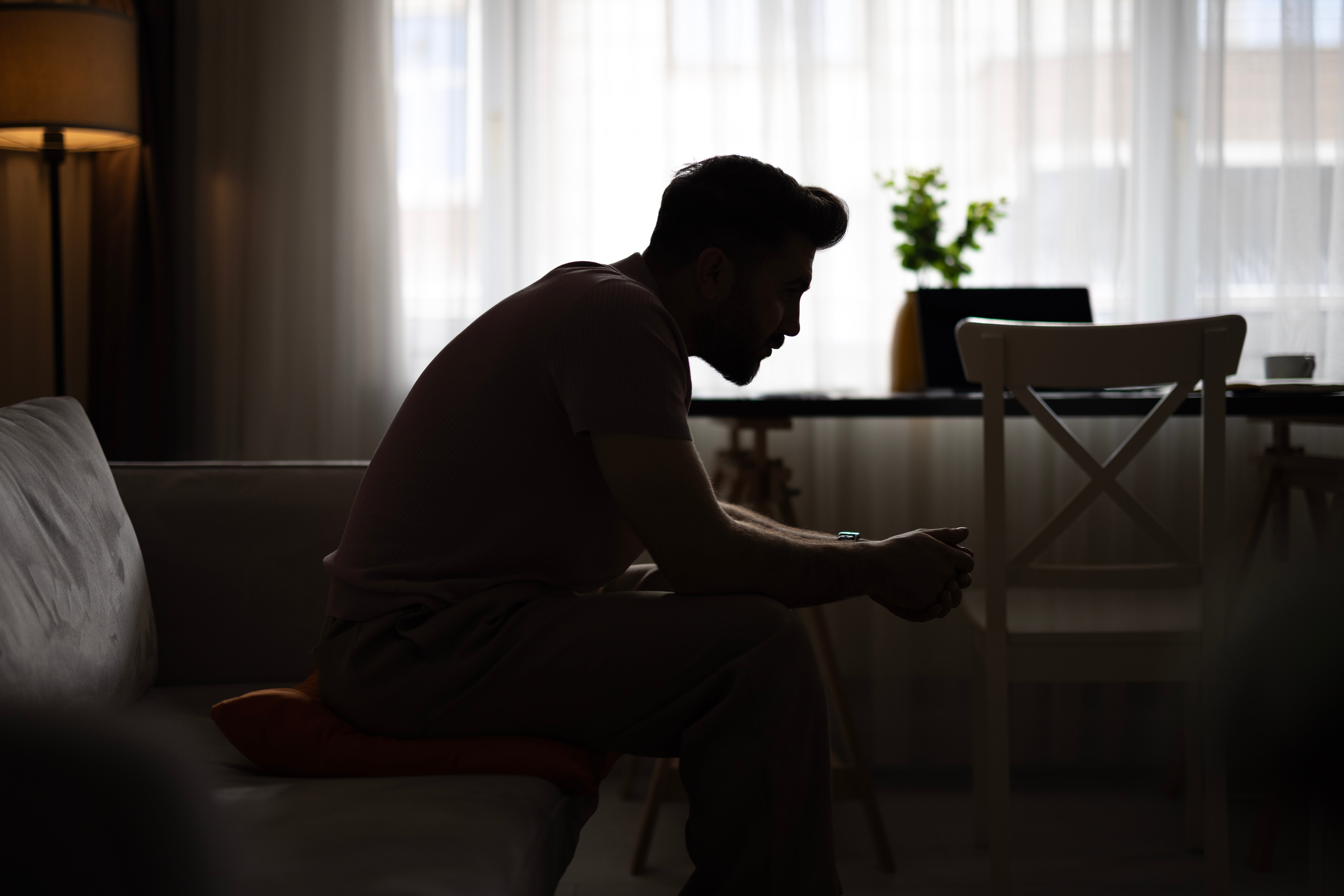 Silhouette of a person sitting on a couch, looking contemplative, in a softly lit room with a desk and chair in the background