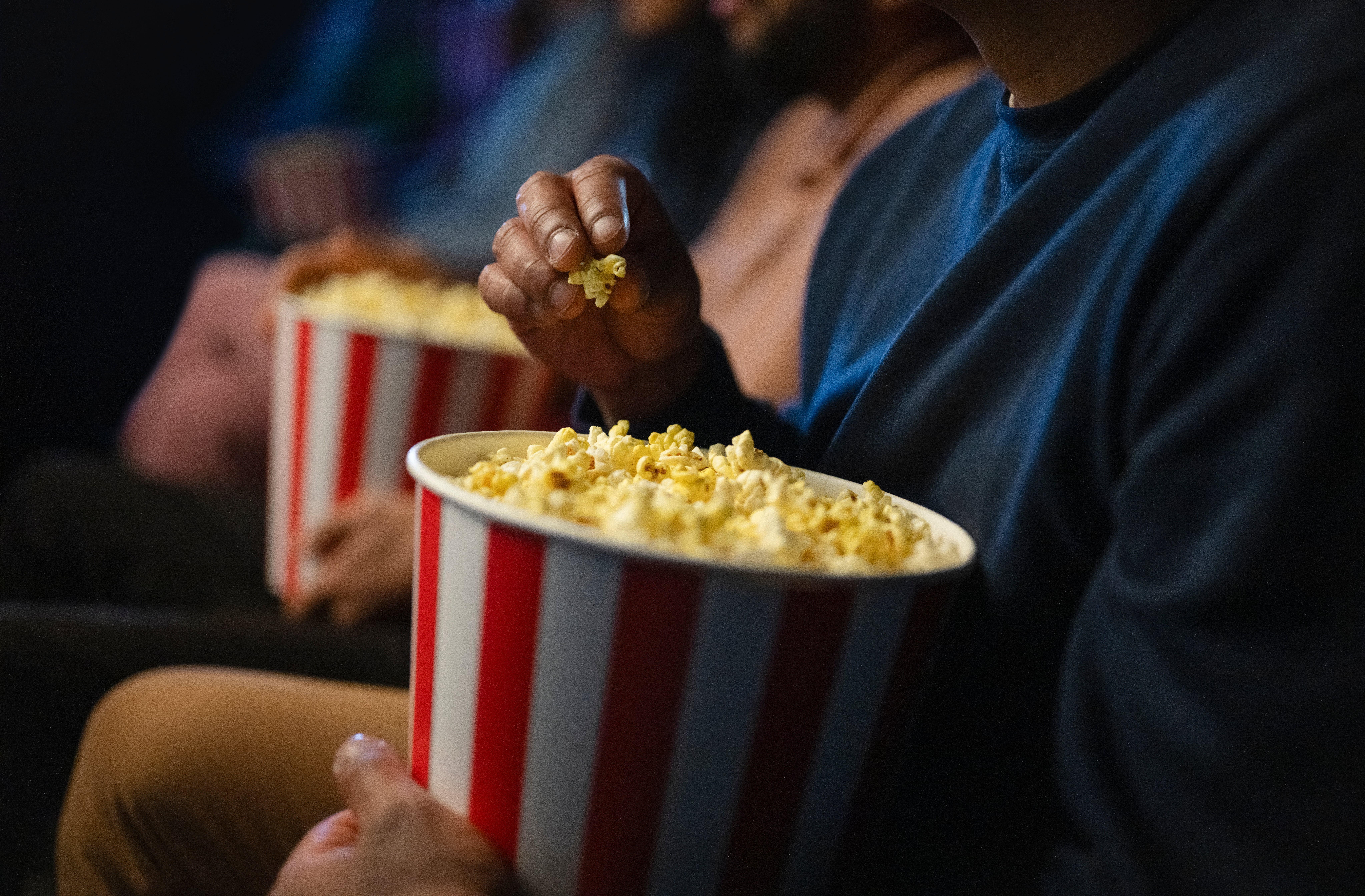 Orang-orang yang duduk di teater, masing-masing memegang wadah popcorn bergaris besar, fokus pada layar