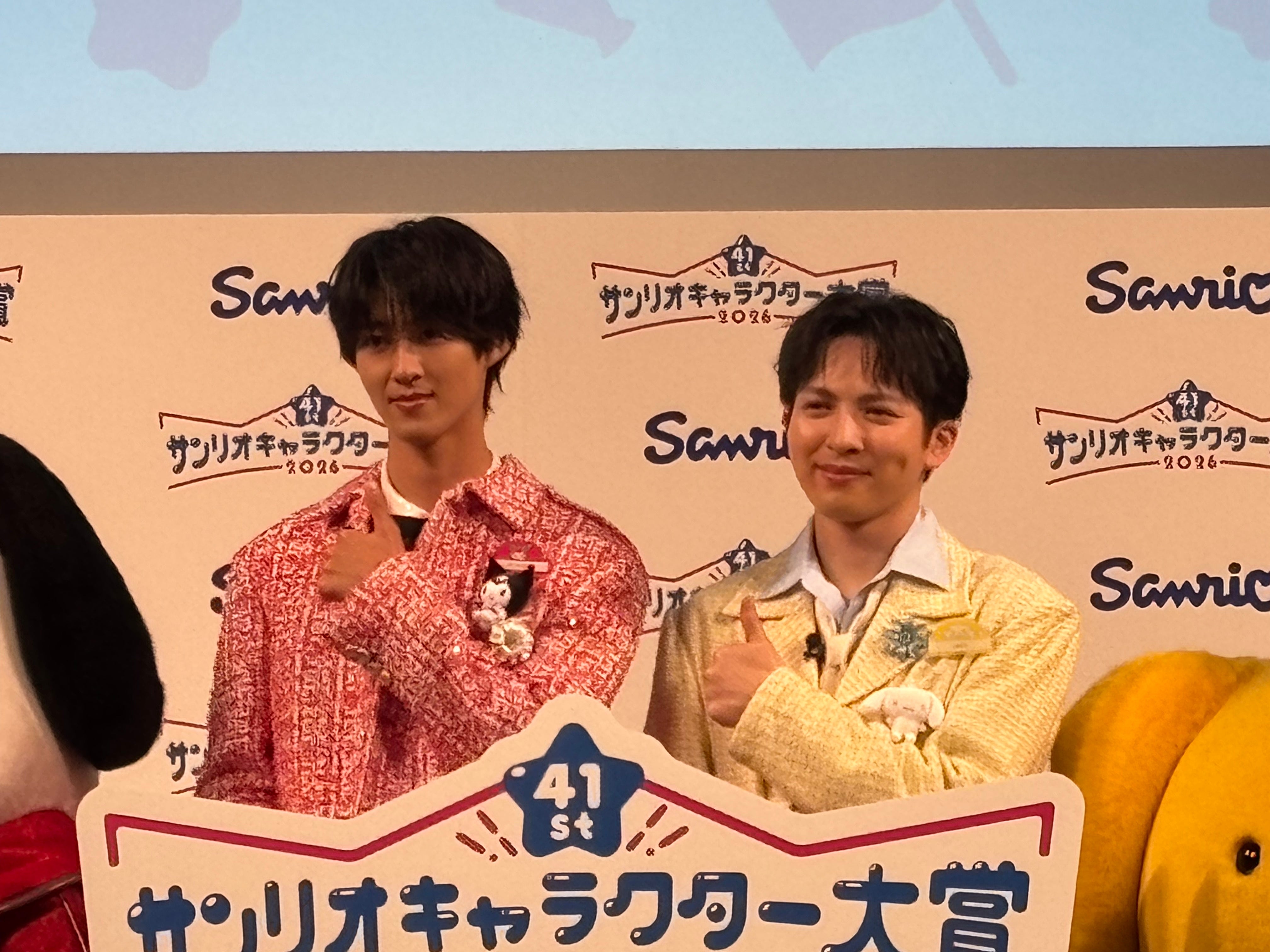 男性2人がサンリオキャラクター大賞の看板前でポーズ。左はカジュアルな赤のニット、右はフォーマルな黄色のジャケット。