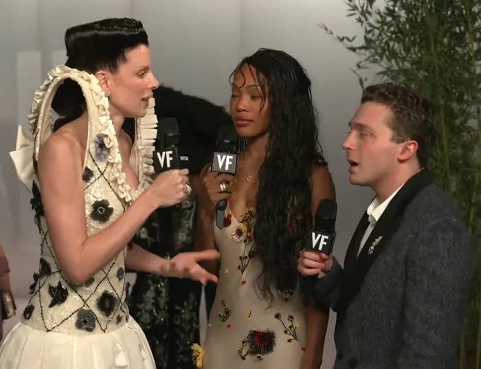 Three radical   clasp  microphones connected  a reddish  carpet event. One wears an ornate formal  with ample  shoulders, different  a formal  with angiosperm  details, and the 3rd  a suit