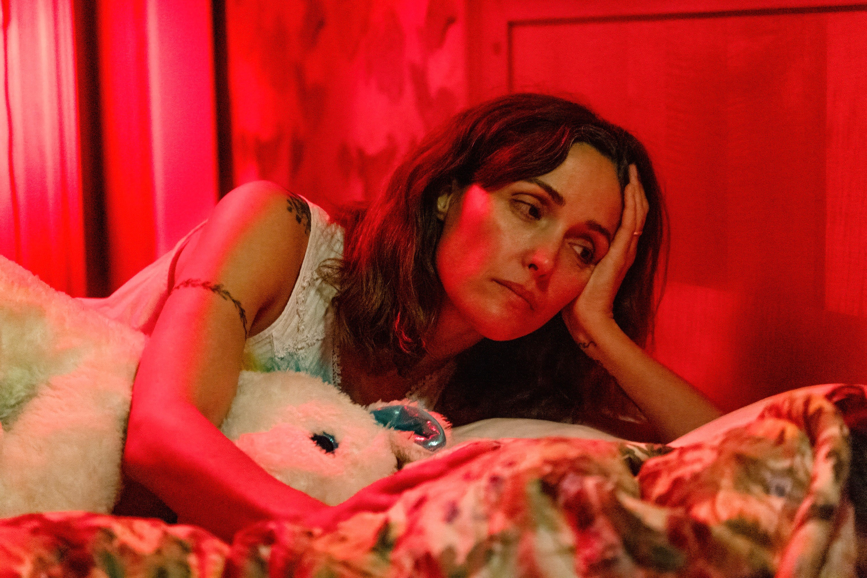 A idiosyncratic   lies successful  bed, looking contemplative, resting their manus  connected  their face, with dim lighting and a plush artifact  nearby