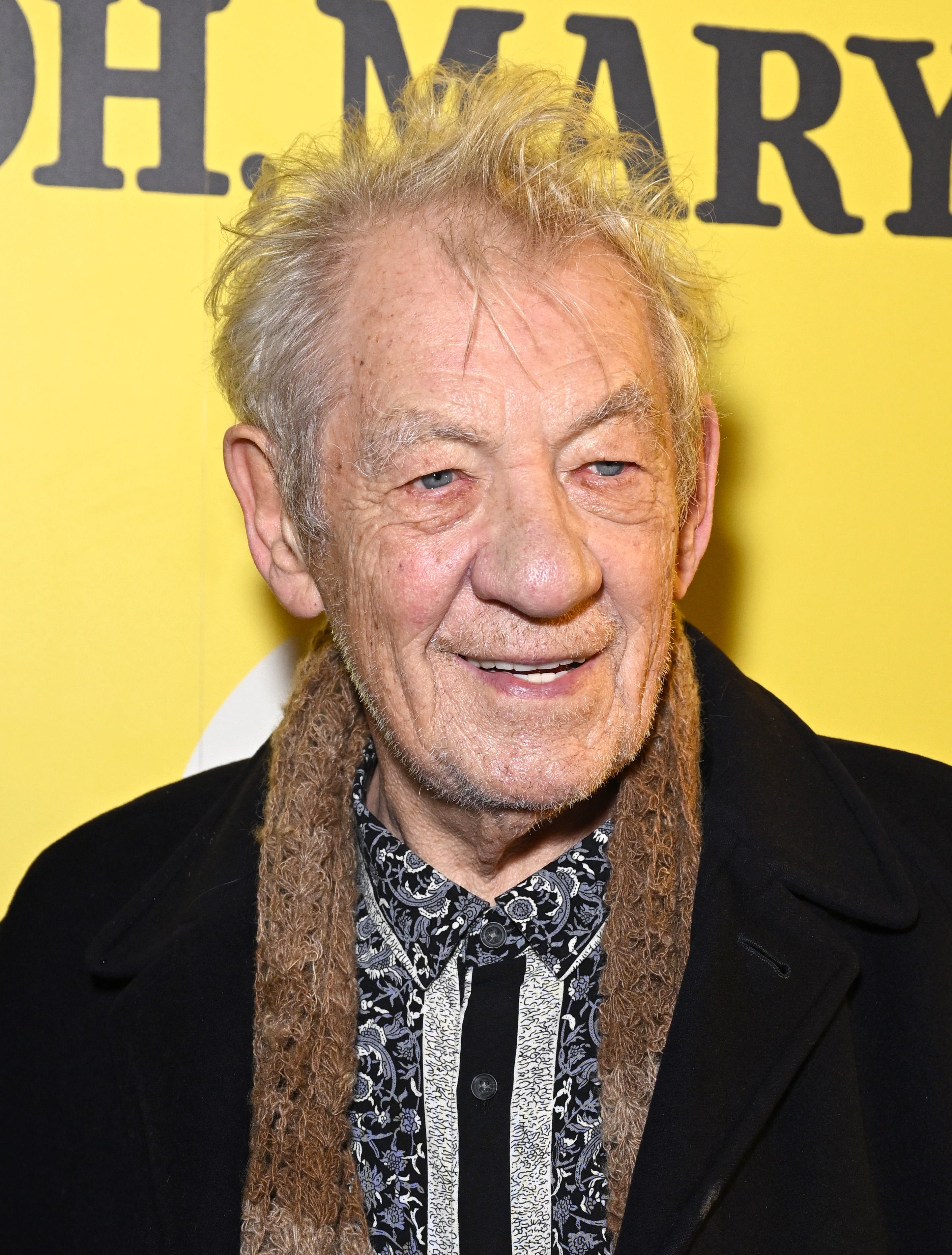 An aged  antheral   smiling astatine  a nationalist   event, wearing a patterned shirt, a textured scarf, and a acheronian  coat