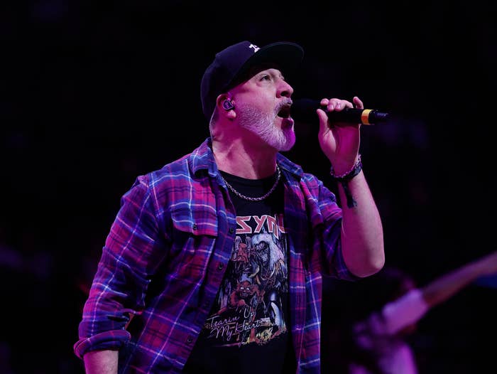 A antheral   performing connected  stage, wearing a flannel garment  implicit    a graphic tee and a cap, singing into a microphone