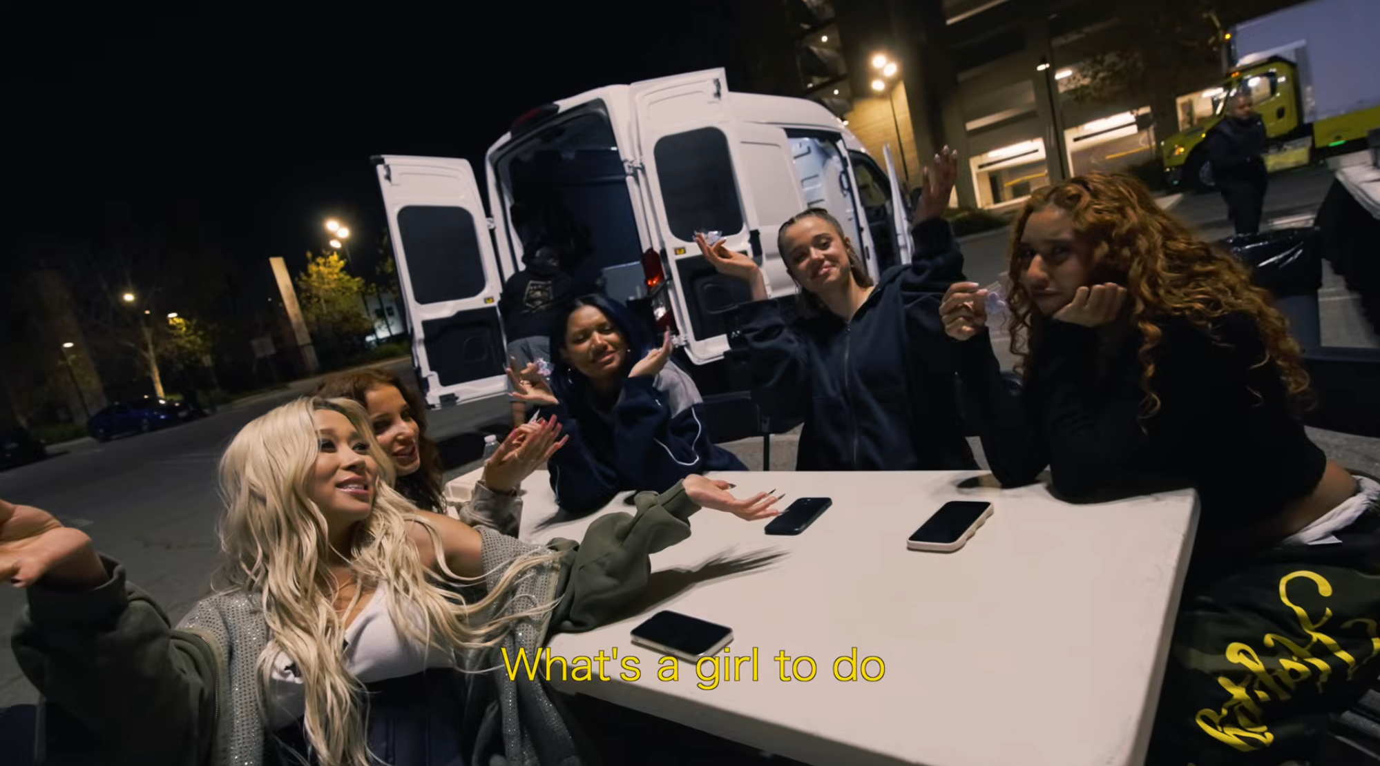 Group of five people seated at a table outside at night, smiling and posing with open arms, text reads: "What's a girl to do"