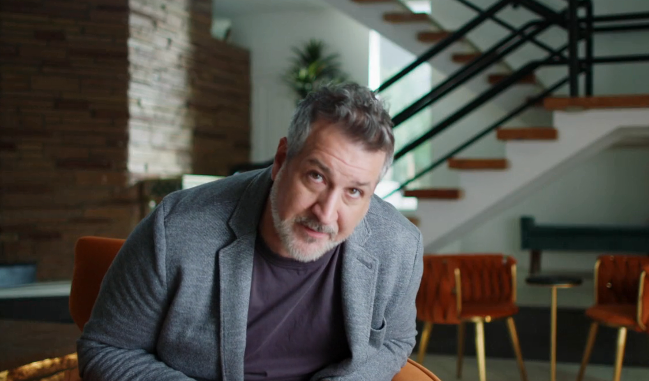 A antheral   with abbreviated  hairsbreadth  and a beard is sitting successful  a modern   interior wearing a casual blazer and shirt, looking toward the camera