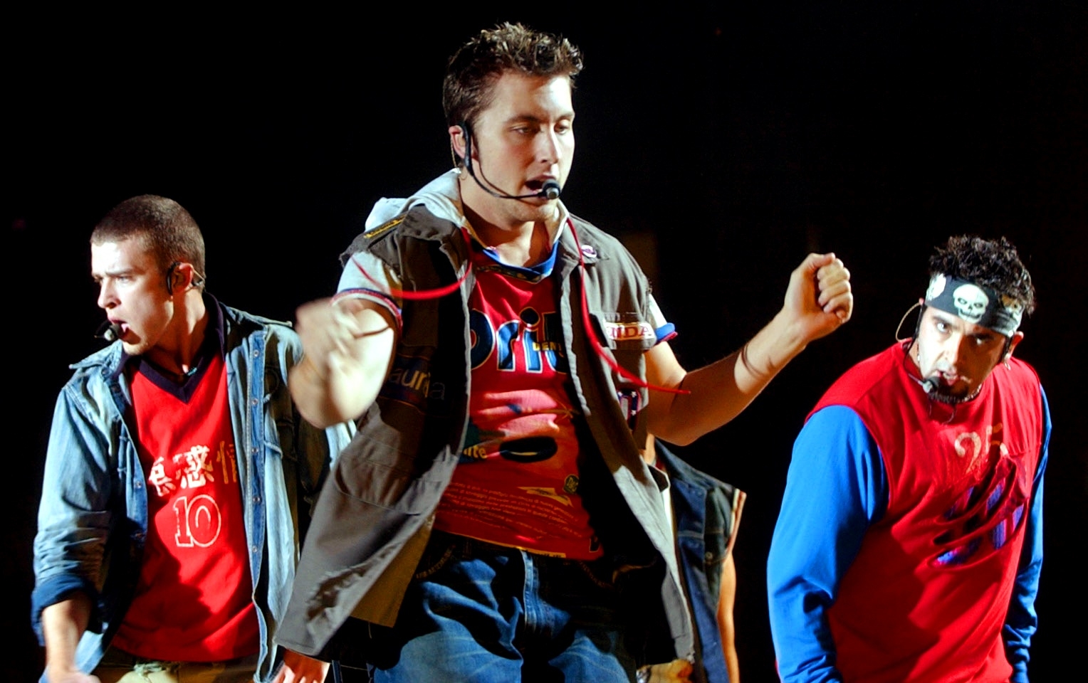 Three performers connected  stage, wearing casual layered outfits, each   with a microphone headset, actively dancing during a concert