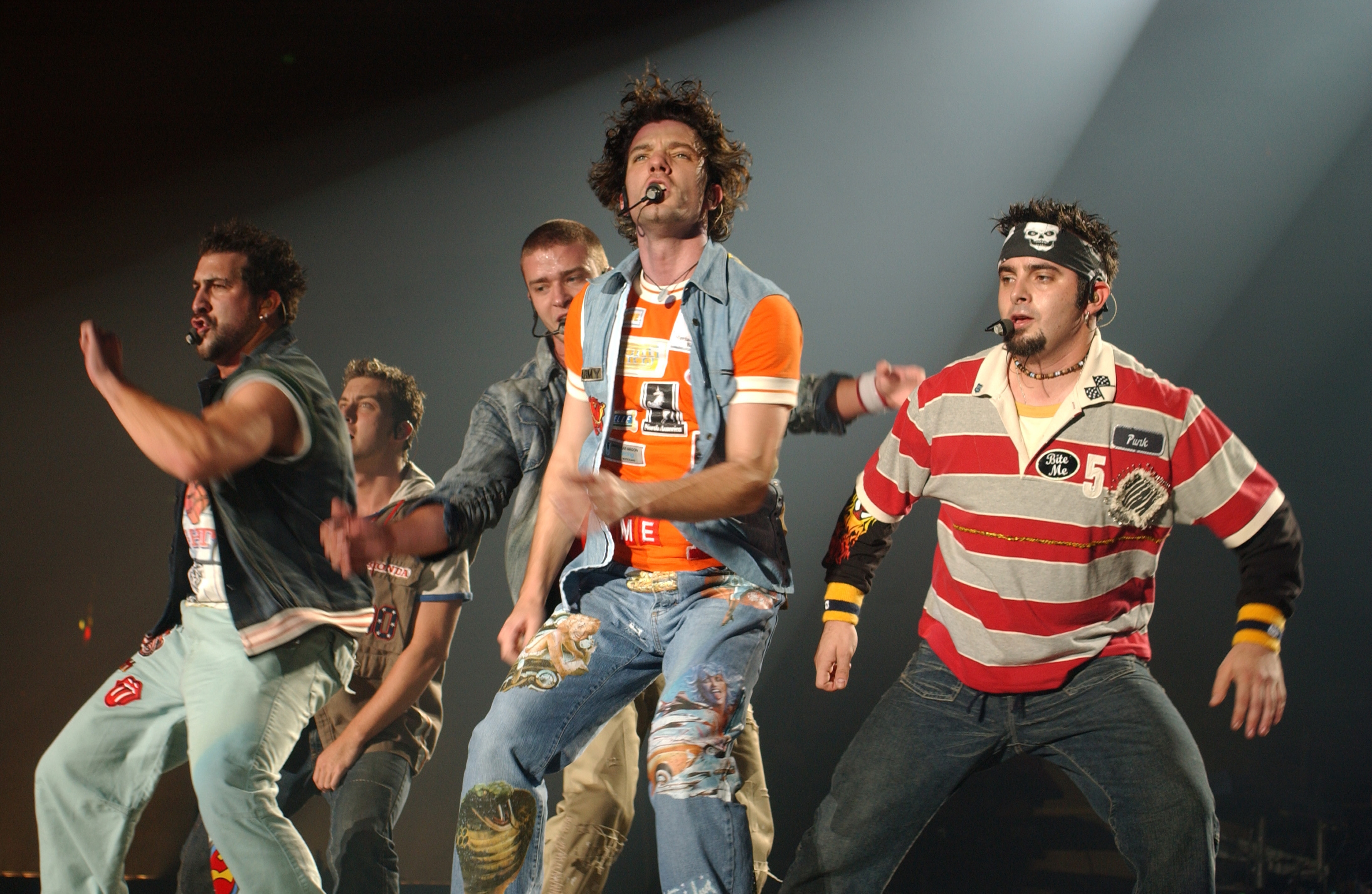 Five performers connected  stage, mid-dance, wearing casual, eclectic outfits with microphones