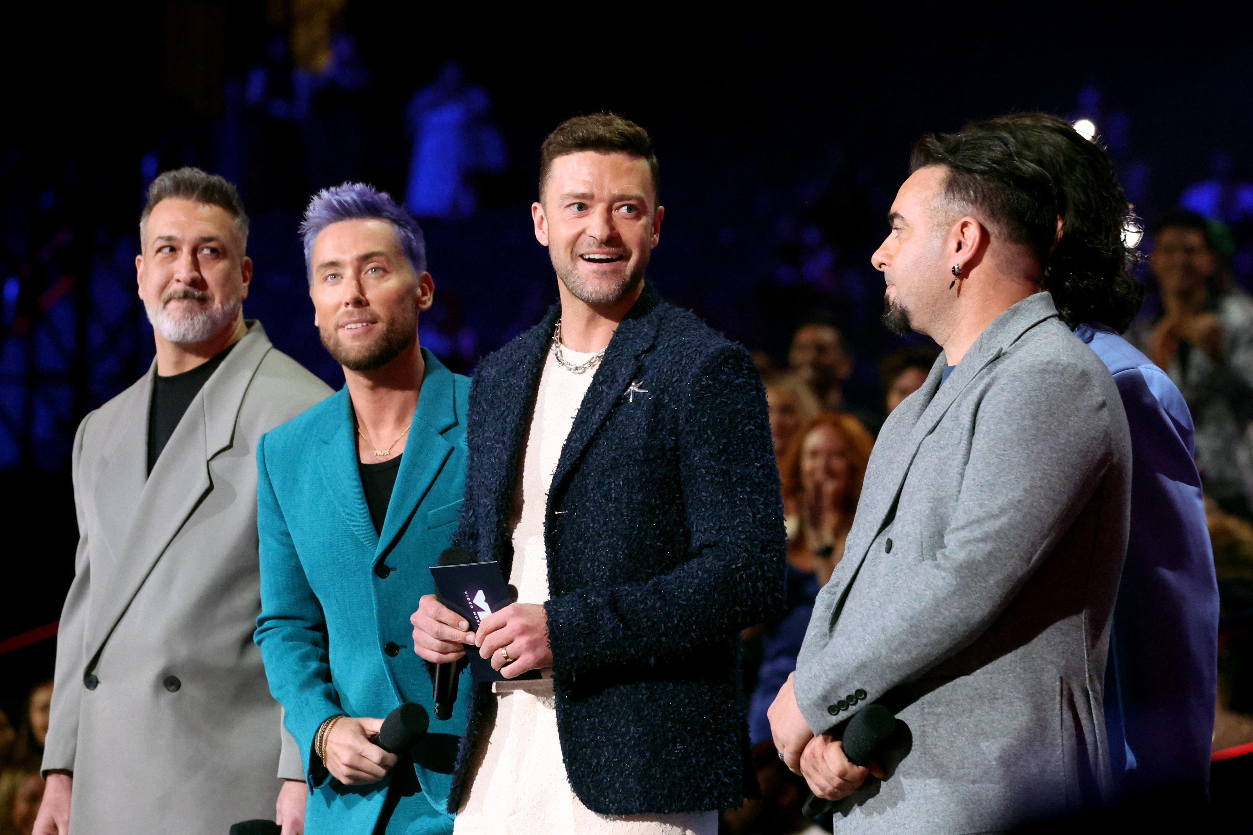 A radical  of 4  radical   successful  stylish suits smiling astatine  an lawsuit   stage, holding microphones, with a lively assemblage  successful  the background