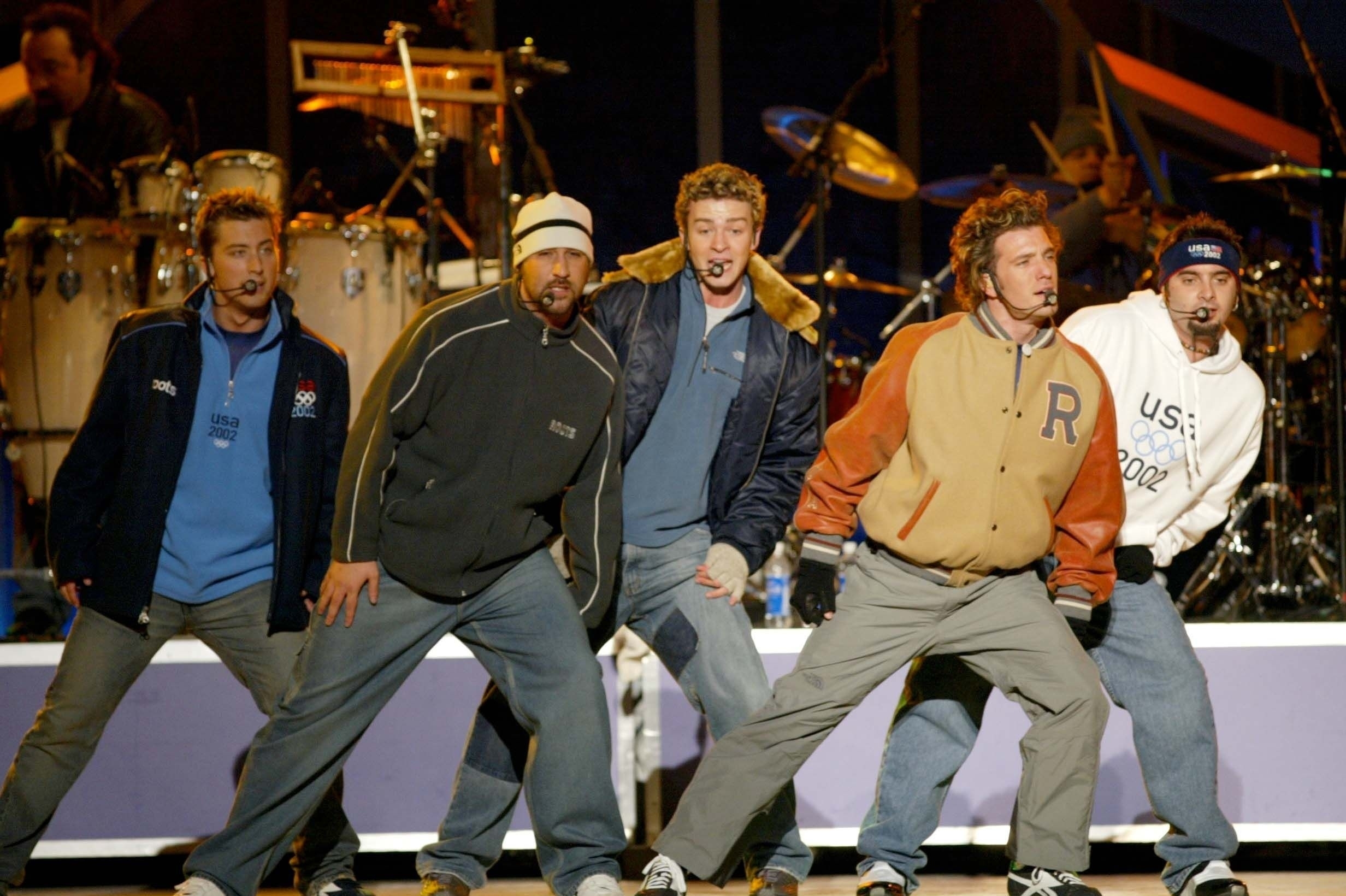 A radical  of antheral  performers sing and creation   connected  stage, wearing casual and sporty outfits, with a drum acceptable   successful  the background