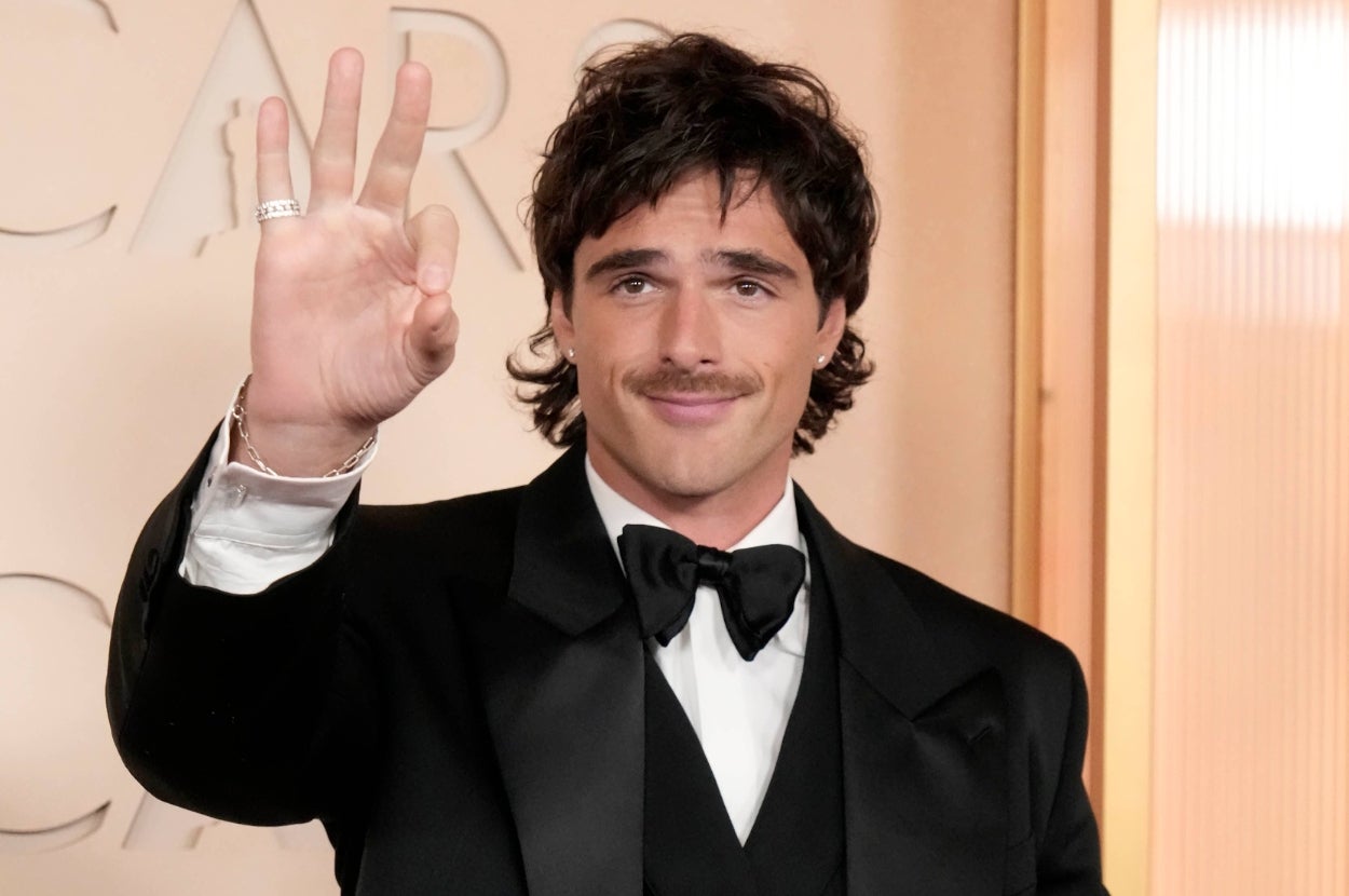 A person on the red carpet wearing a black tuxedo and bow tie, gesturing with one hand. Background features the word "Oscars."