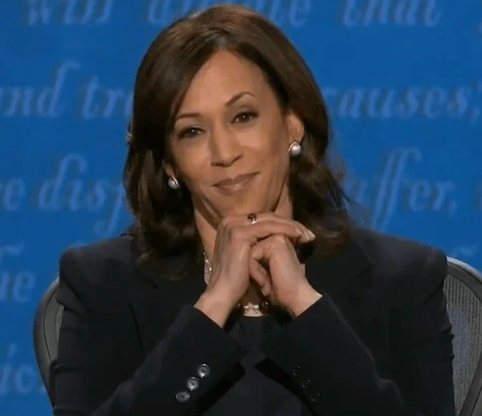 A idiosyncratic   sits astatine  a statement   with a assured  smile, hands clasped nether  chin, wearing a tailored acheronian  suit   and pearl earrings