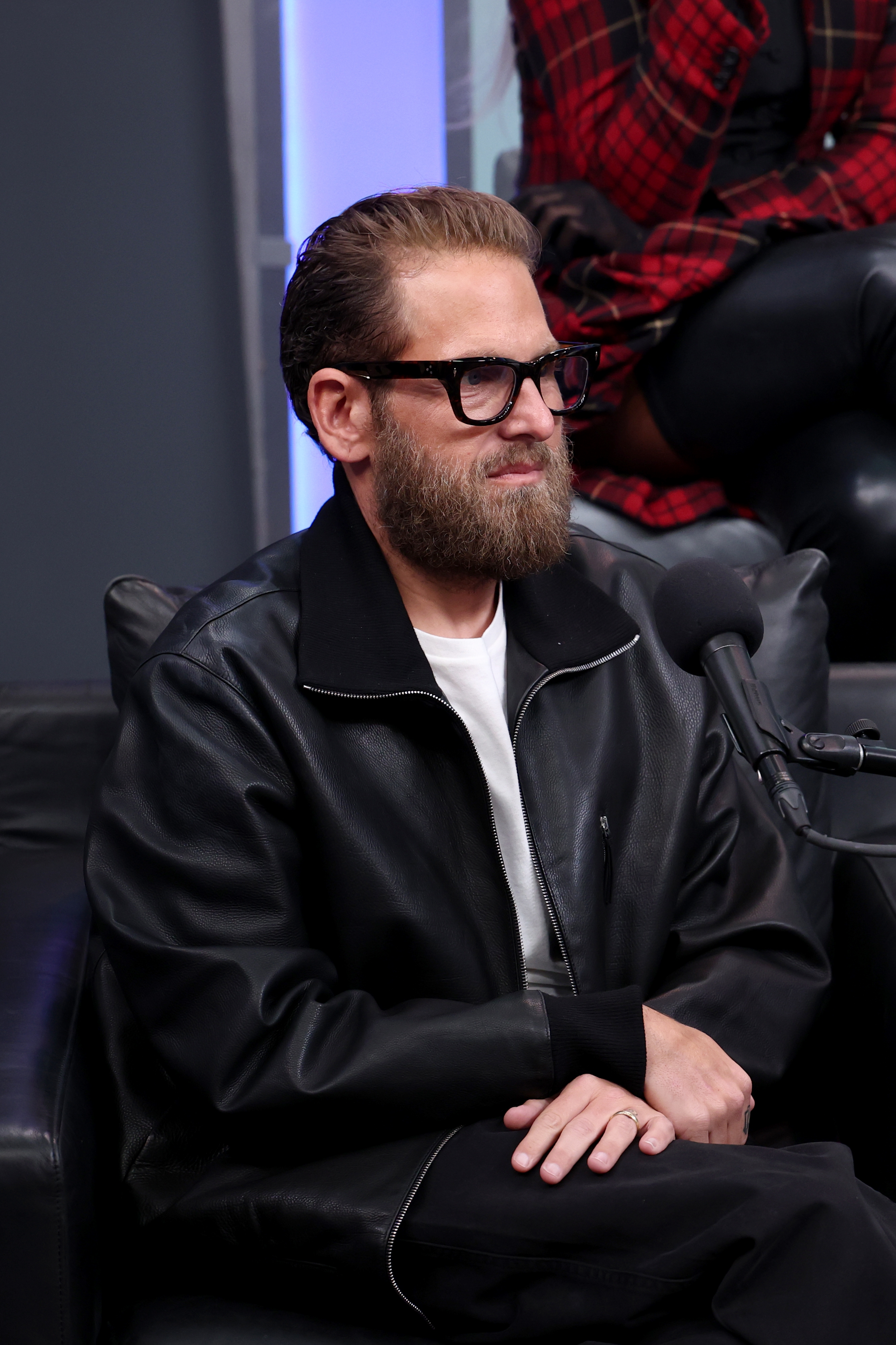 A idiosyncratic with glasses and a beard sits connected a achromatic seat wearing a achromatic leather overgarment and achromatic shirt, speaking into a microphone