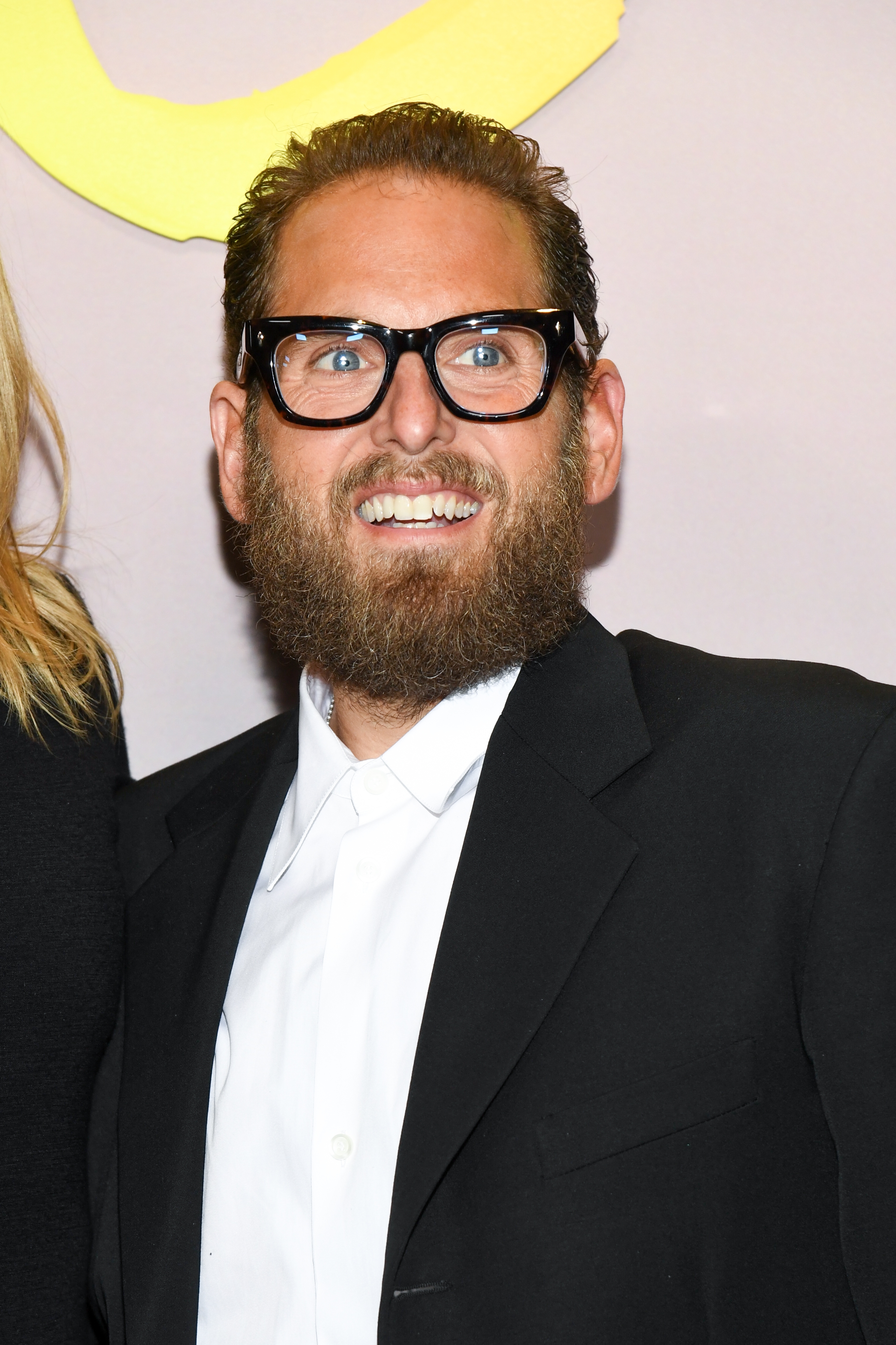 A idiosyncratic successful a suit and achromatic garment is smiling astatine an event, wearing thick-framed glasses and sporting a afloat beard