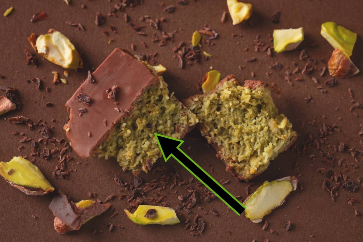 Chocolate-covered pistachio-flavored cake, partially sliced, sprinkled with chocolate shavings and pistachio pieces on a brown surface