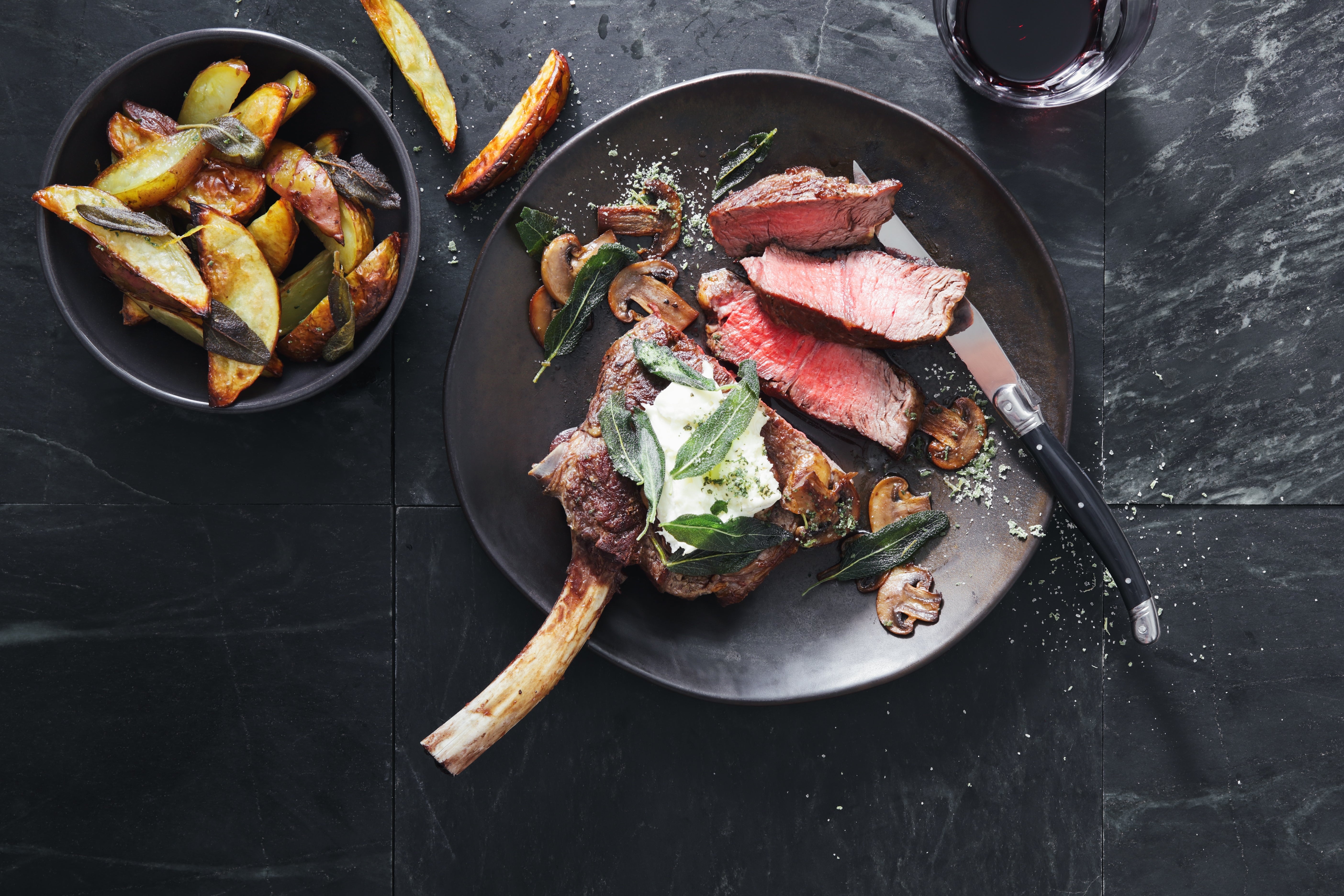 A sheet  with sliced steak and grilled vegetables, including mushrooms, adjacent  to a vessel  of roasted potatoes. A weapon  rests beside the steak