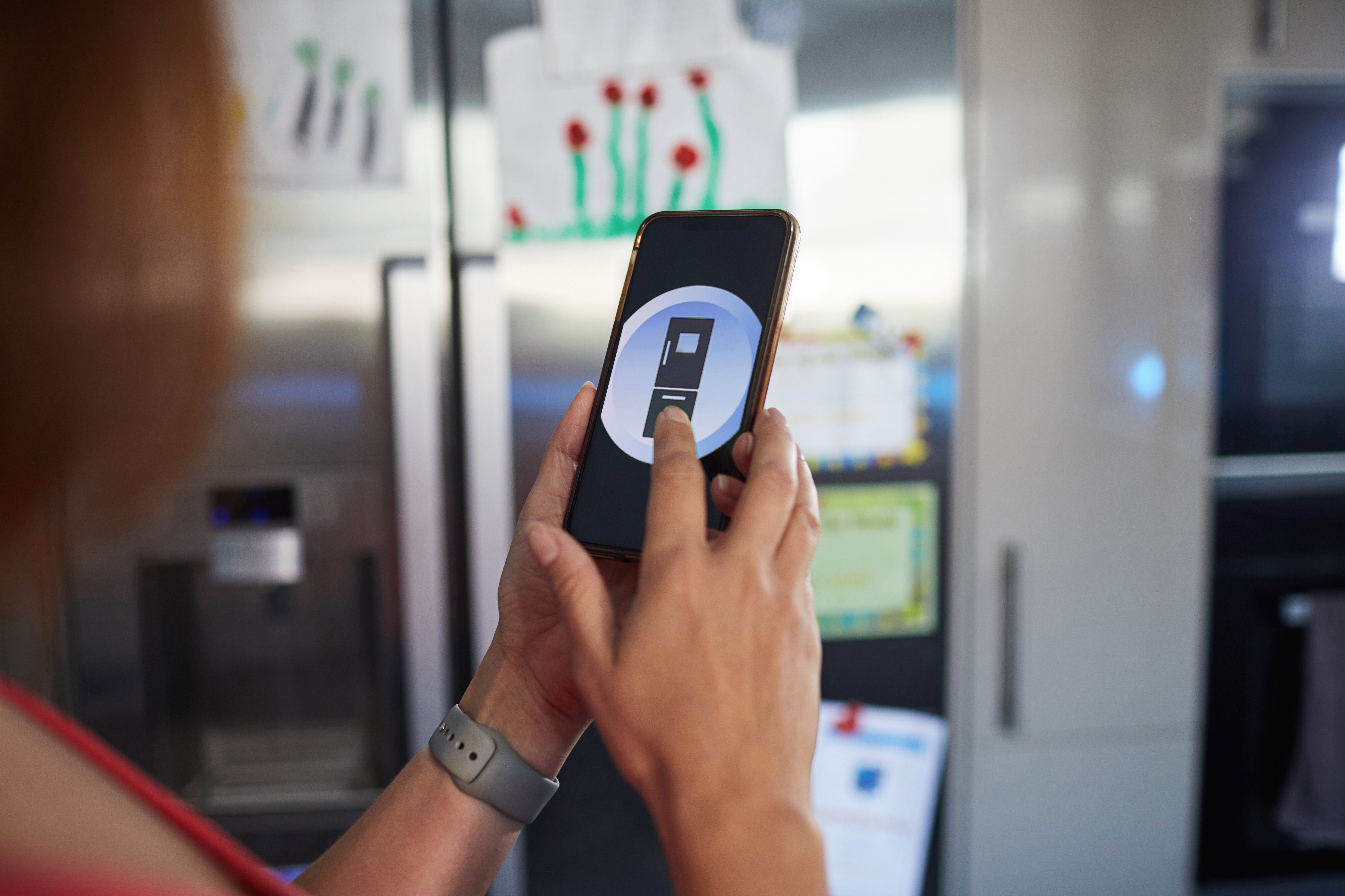Person holding a smartphone displaying a astute  location  app interface, lasting  successful  beforehand   of a refrigerator with children's artwork