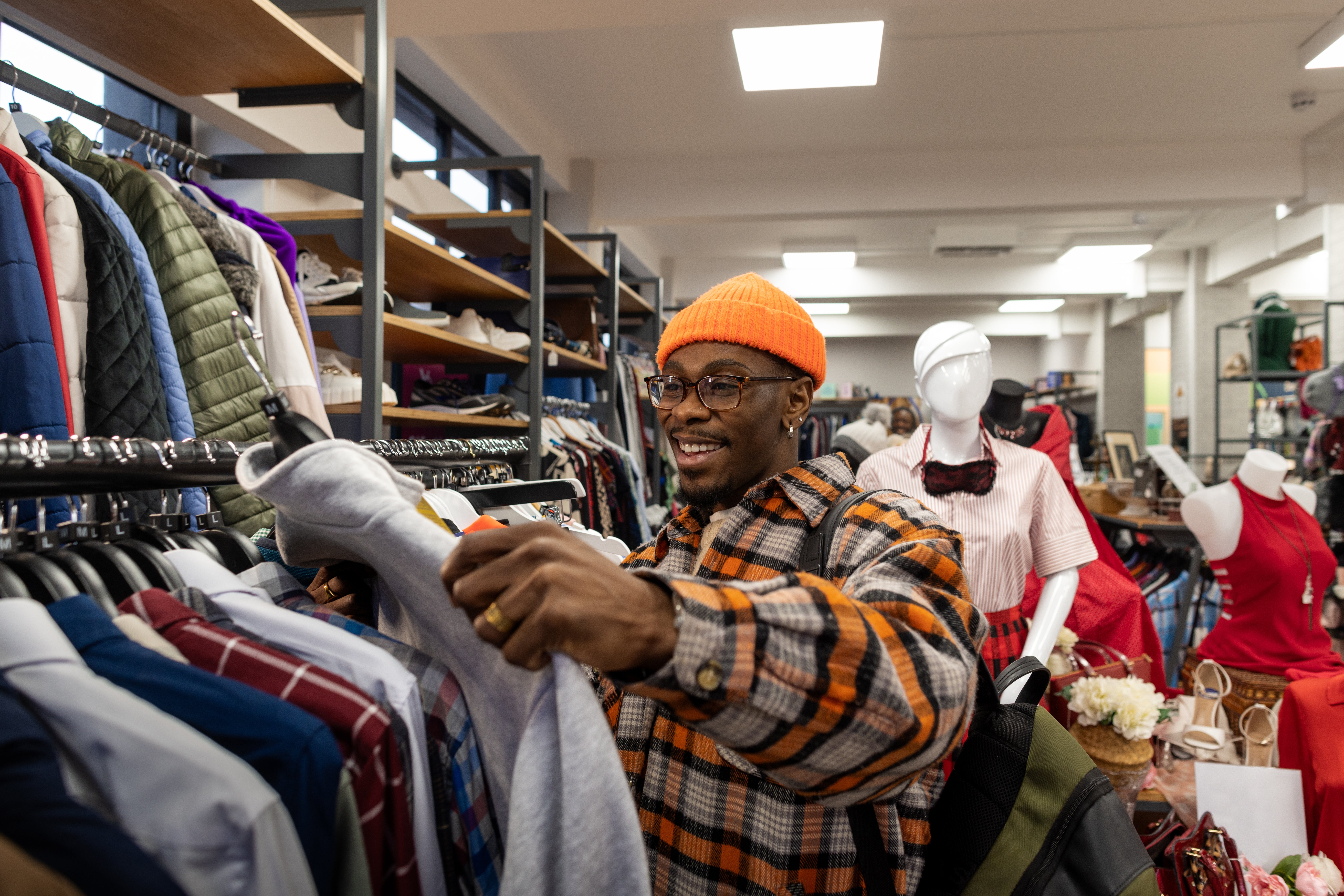 A idiosyncratic   successful  a plaid garment  and orangish  beanie happily browses apparel  successful  a store, holding a grey  sweatshirt, with mannequins successful  the background