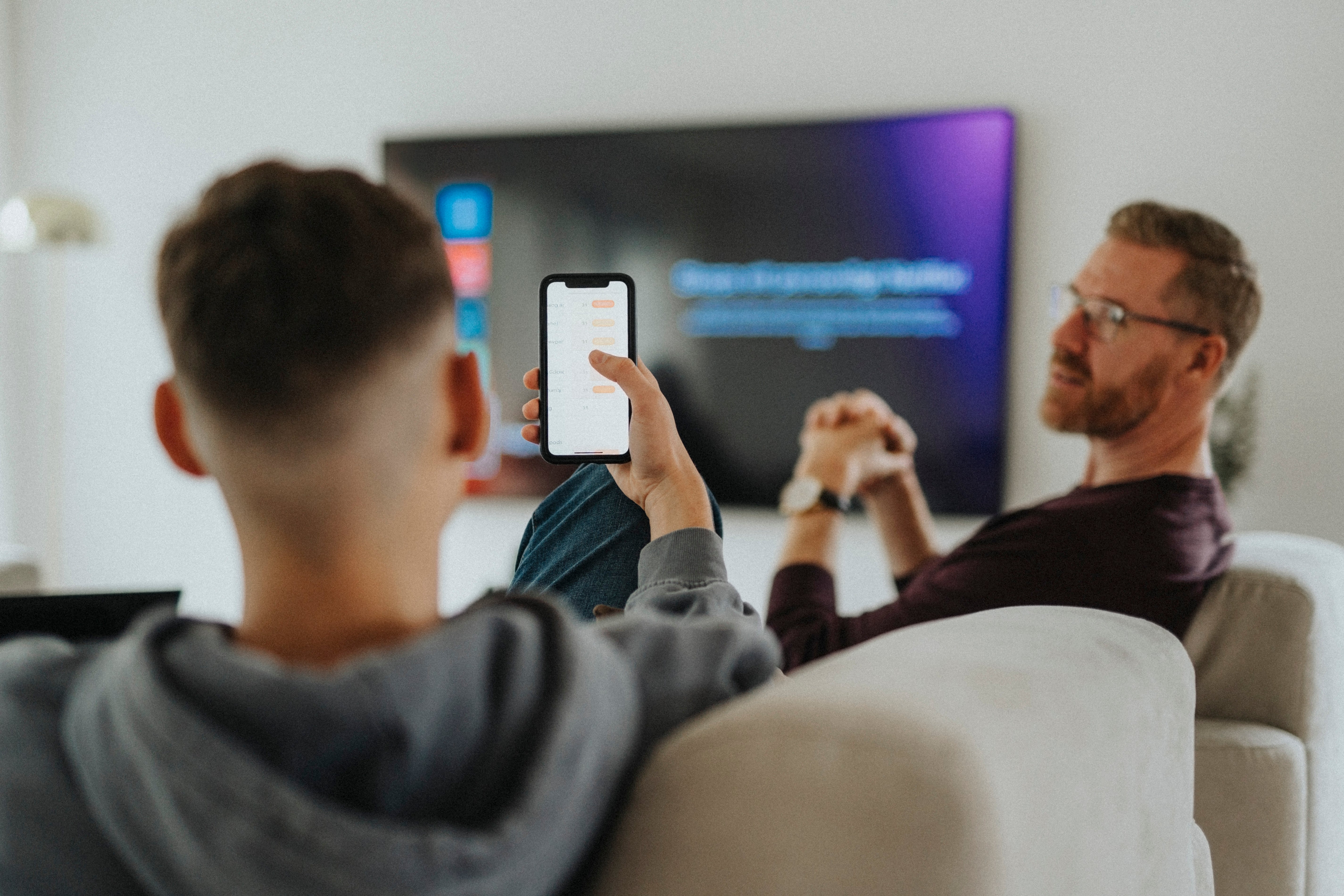 Two radical   connected  a couch; 1  holds a telephone  up, showing the surface  to the other. A TV is successful  the background