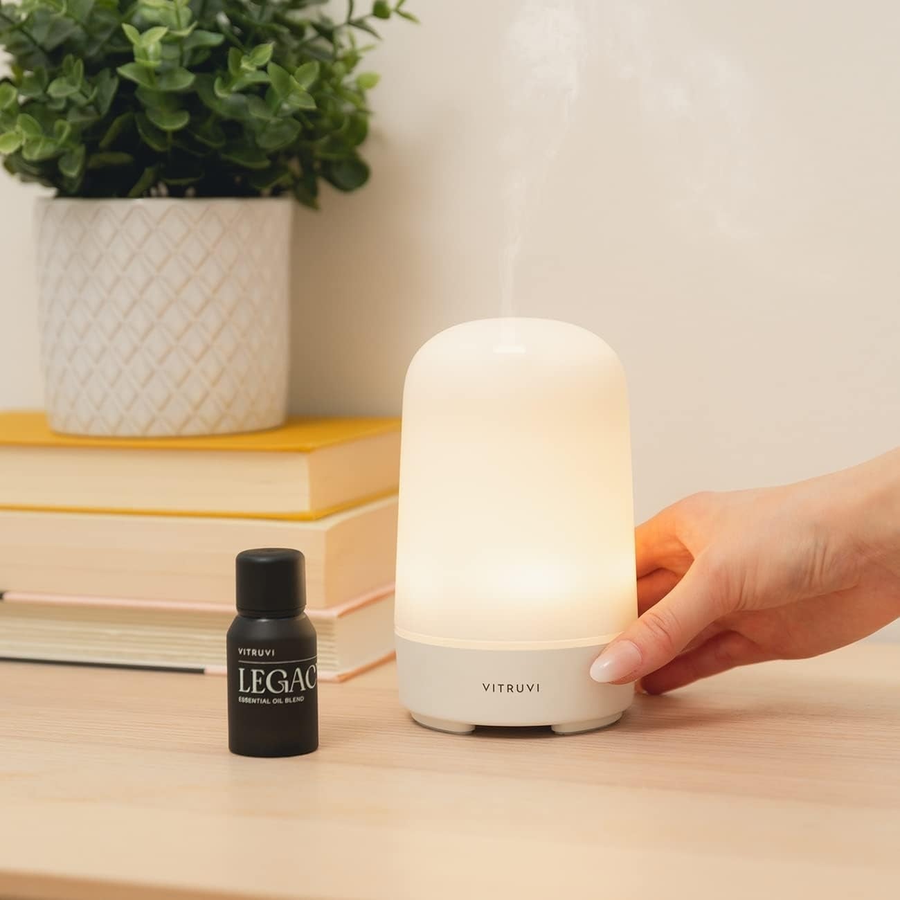 Hand adjusting a sleek, minimalist essential oil diffuser next to a LEGACY oil bottle on a wooden table with books and a plant in the background