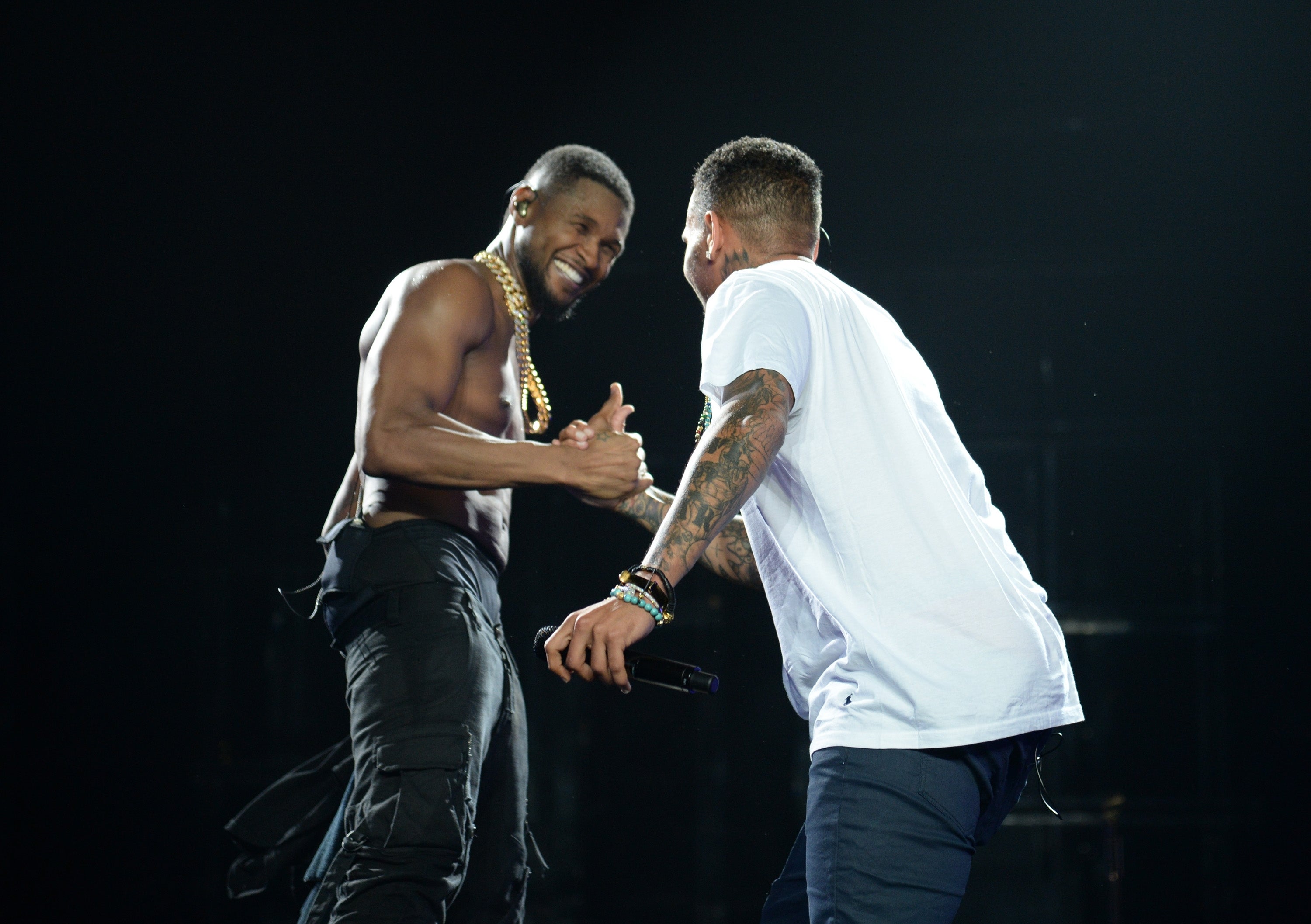 To utøvere deler et vennlig øyeblikk på scenen, utveksler et håndtrykk og smiler. Den ene er skjorteløs og har på seg svarte bukser; den andre har på seg en hvit skjorte