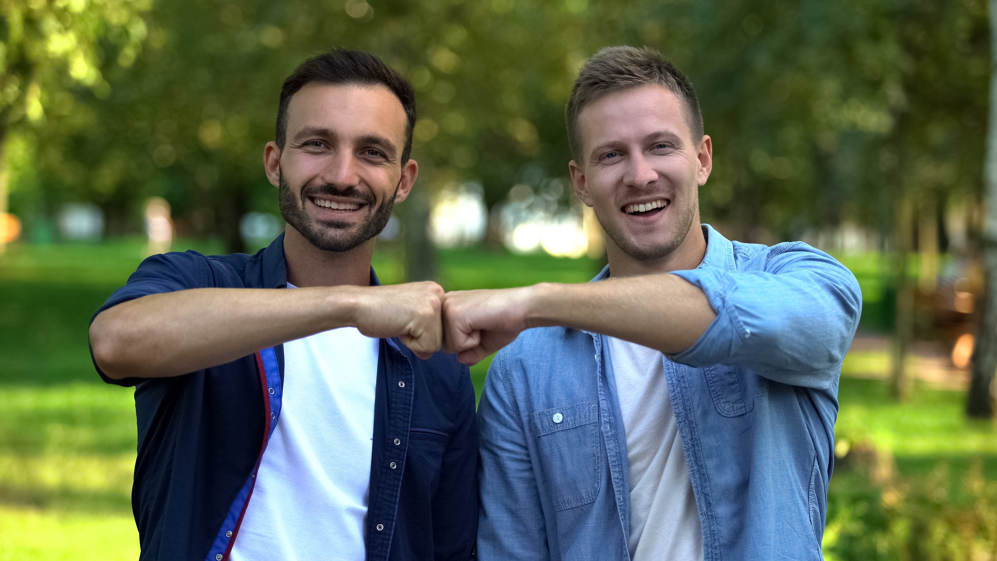 Two people, outdoors, smiling and doing a fist bump. They're casually dressed successful  shirts and T-shirts, enjoying a infinitesimal   together