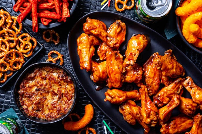 An assortment of snacks including chickenhearted  wings, pretzels, spicy snacks, and dip are laid retired  connected  a textured table, with soda cans nearby