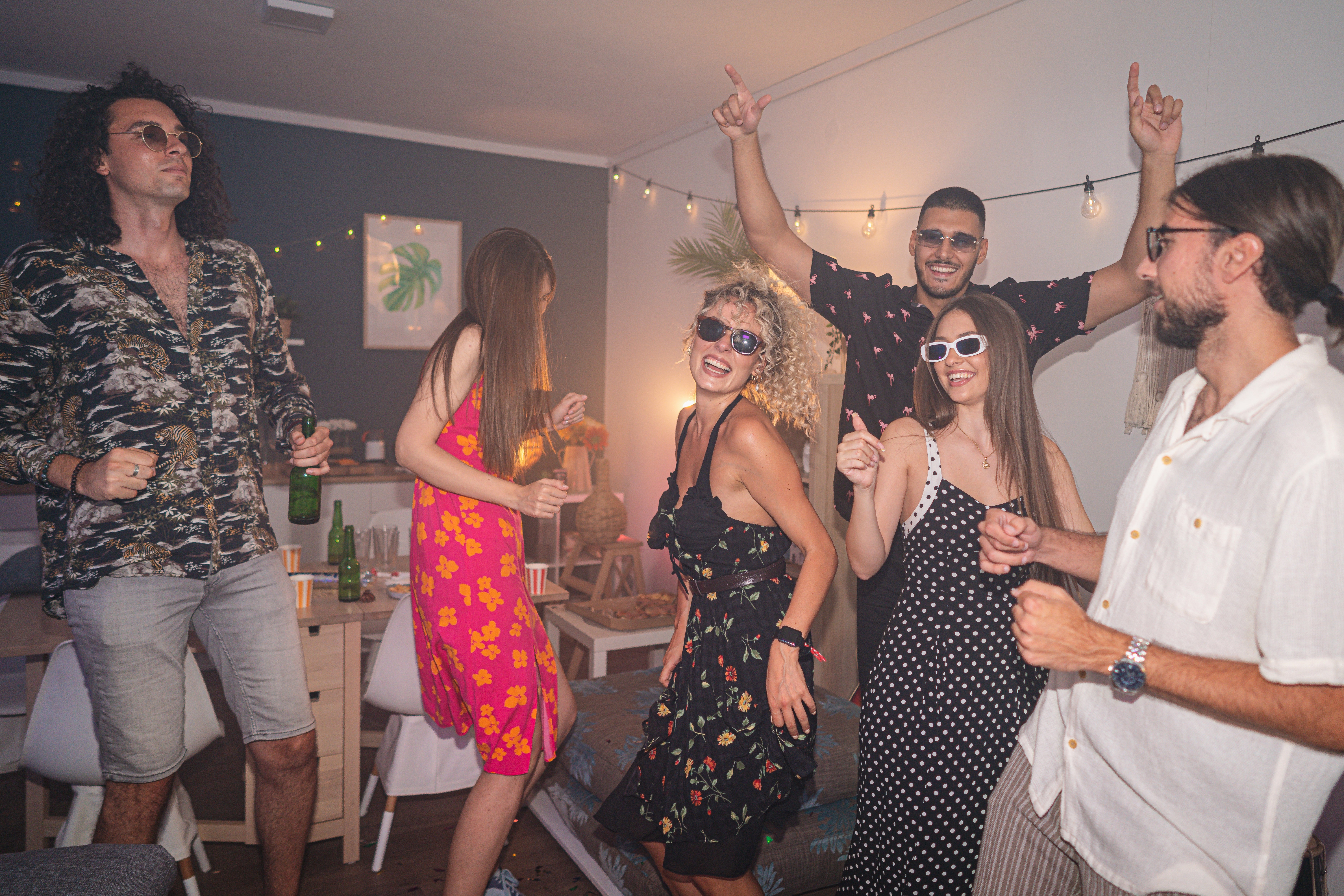 People dancing and enjoying a lively indoor party, wearing casual summertime  outfits and sunglasses, with drawstring  lights and decorations successful  the background