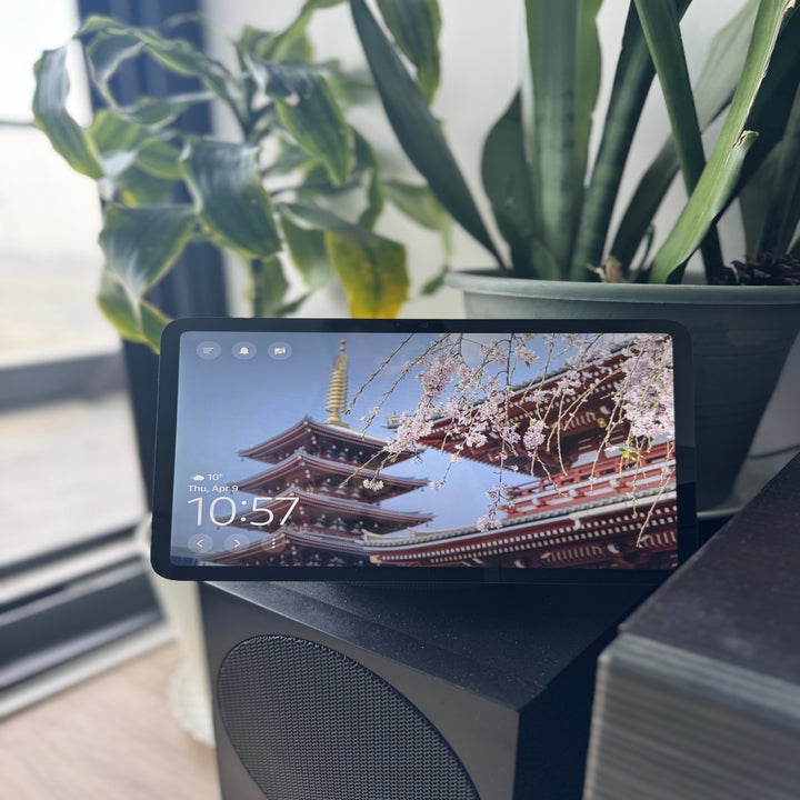 Tablet displaying a scenic pagoda and cherry blossoms on the screen, placed on a speaker beside a large potted plant indoors