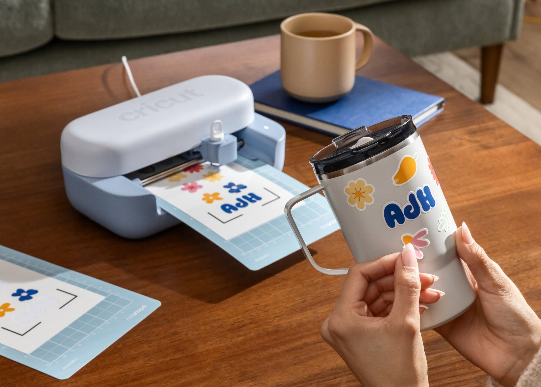 Person placing a personalized sticker with name "Ash" and flowers onto a white mug using a small cutting machine