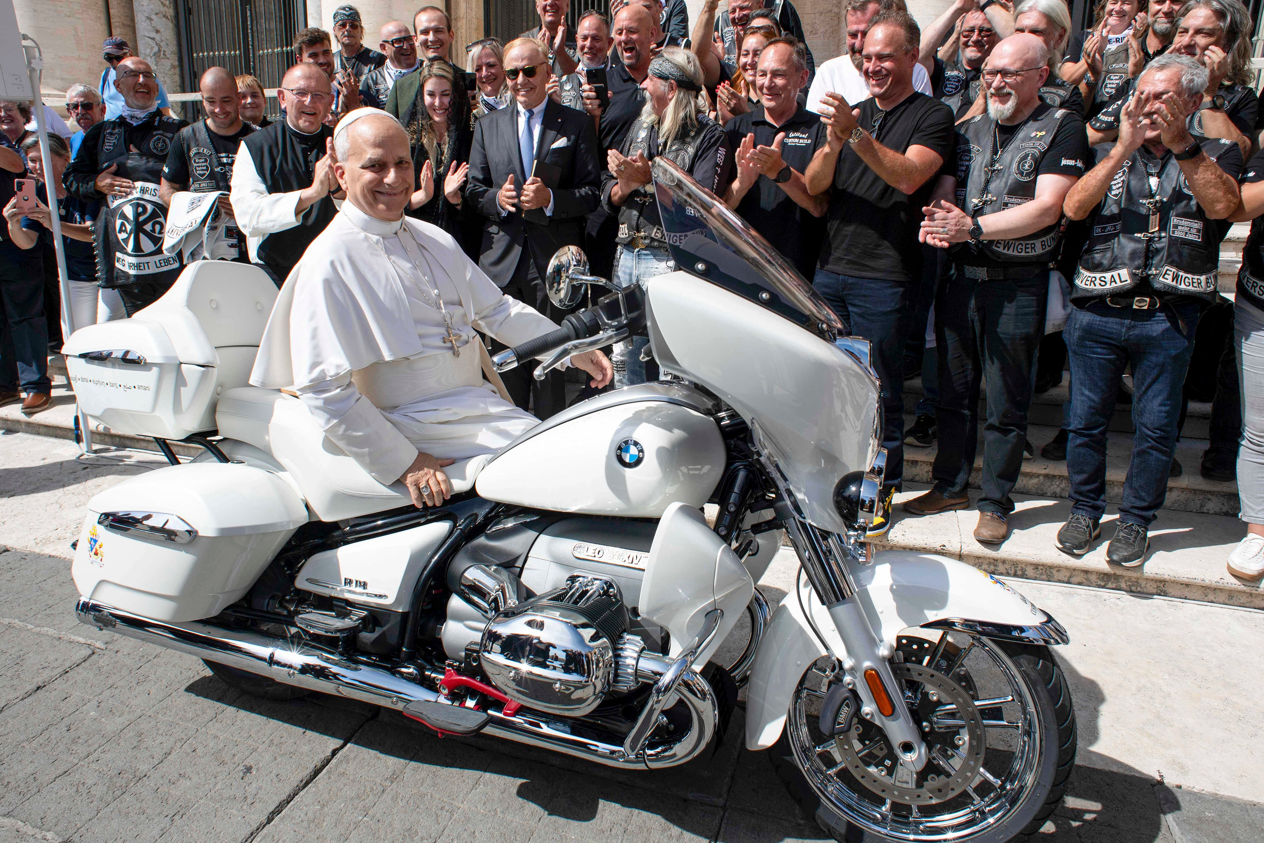 Mann i religiøs antrekk sitter på en hvit motorsykkel, omgitt av applauderende syklister og tilskuere