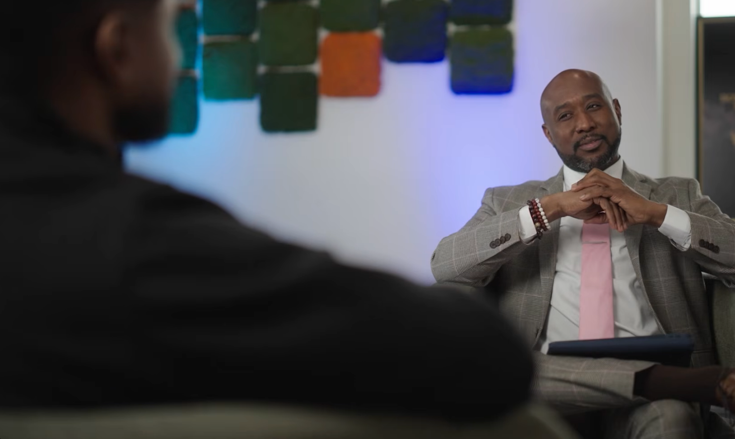 En mann i dress med rosa slips sitter i en intervjusetting og lytter oppmerksomt til en annen person