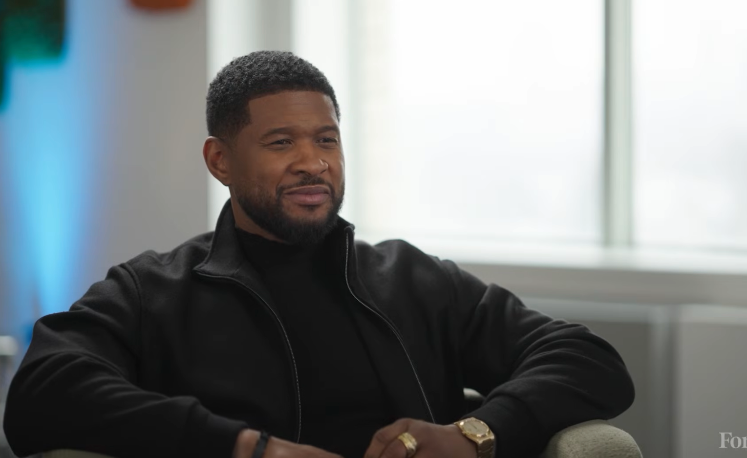 En person i svart rullekrave og jakke sitter i et koselig rom og smiler under et intervju