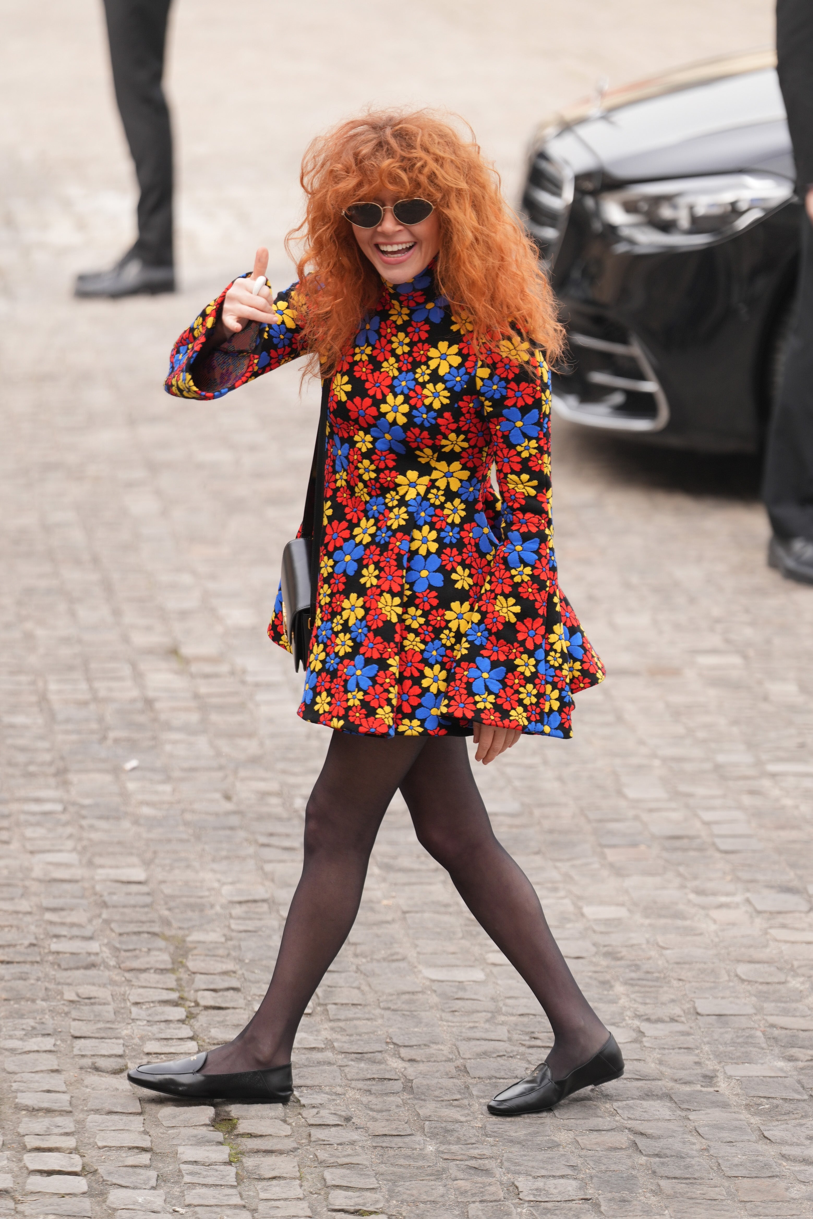 Natasha Lyonne med krøllete hår iført en blomsterkjole og solbriller, gir tommel opp mens hun går på en brosteinssti
