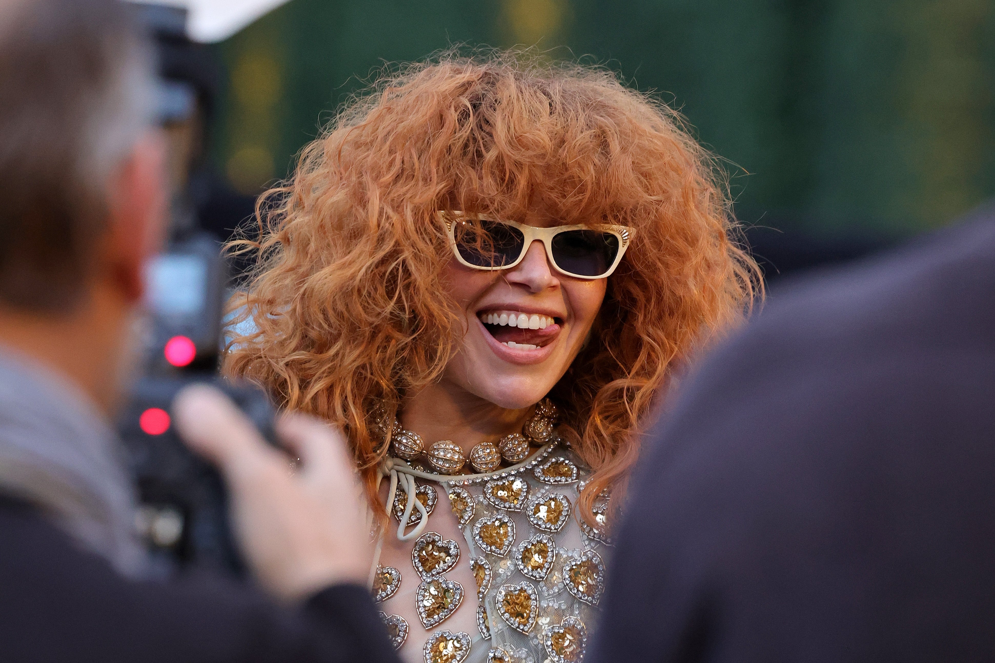 Natasha Lyonne med krøllete hår i solbriller og en pyntet topp smiler