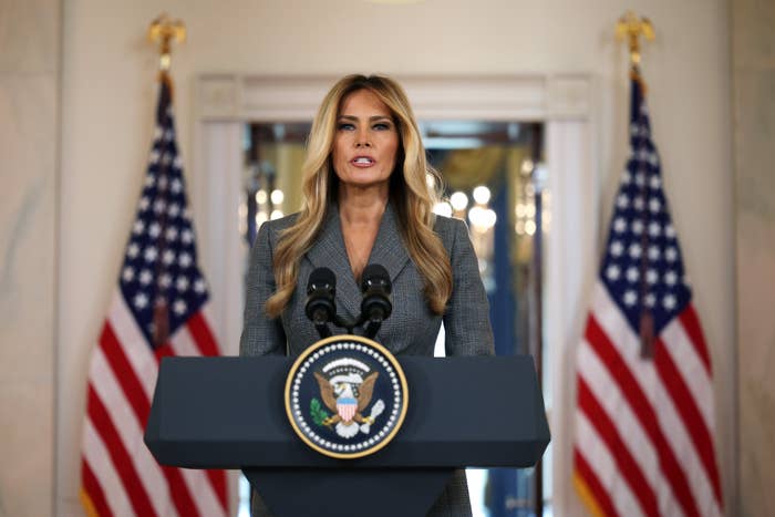 A woman stands at a podium with a U.S. presidential seal, delivering a speech. Two American flags are in the background