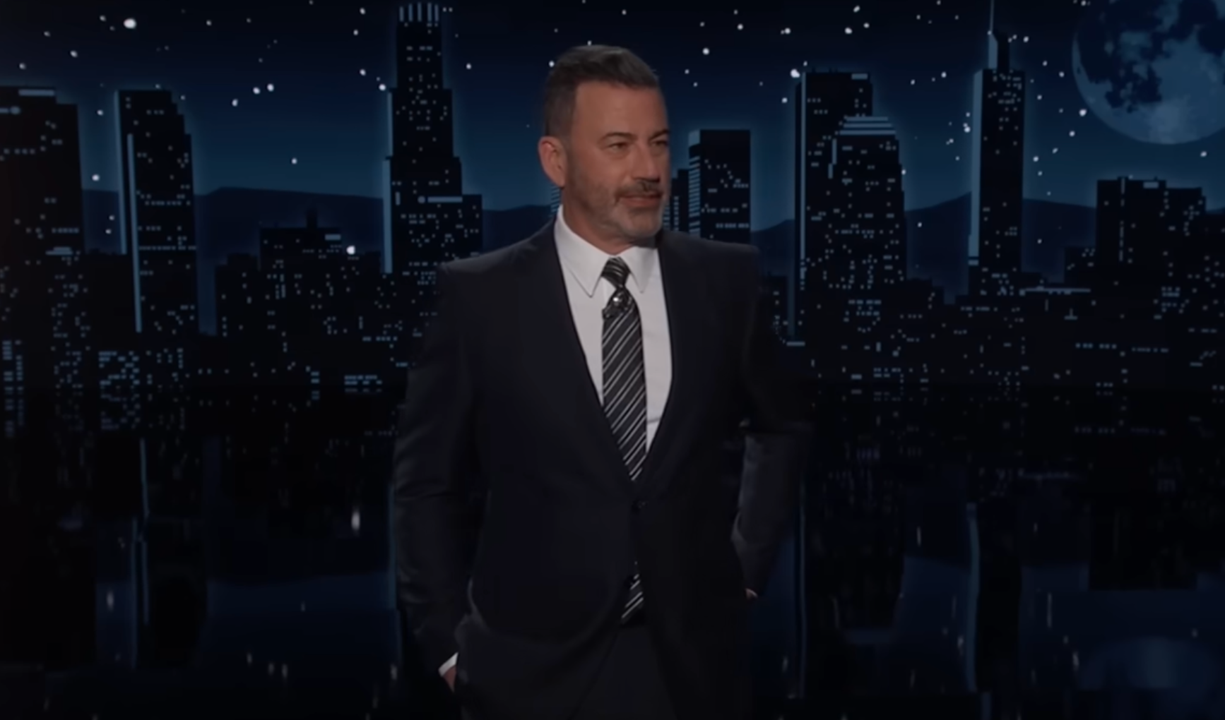 A man in a suit with a striped tie stands on a late-night talk show set against a cityscape backdrop