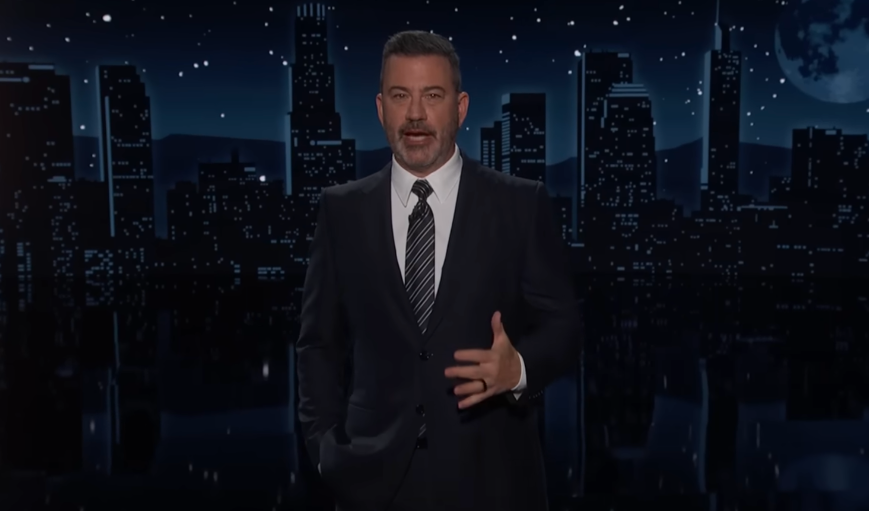 A person in a suit speaks in a cityscape backdrop, likely on a late-night show set
