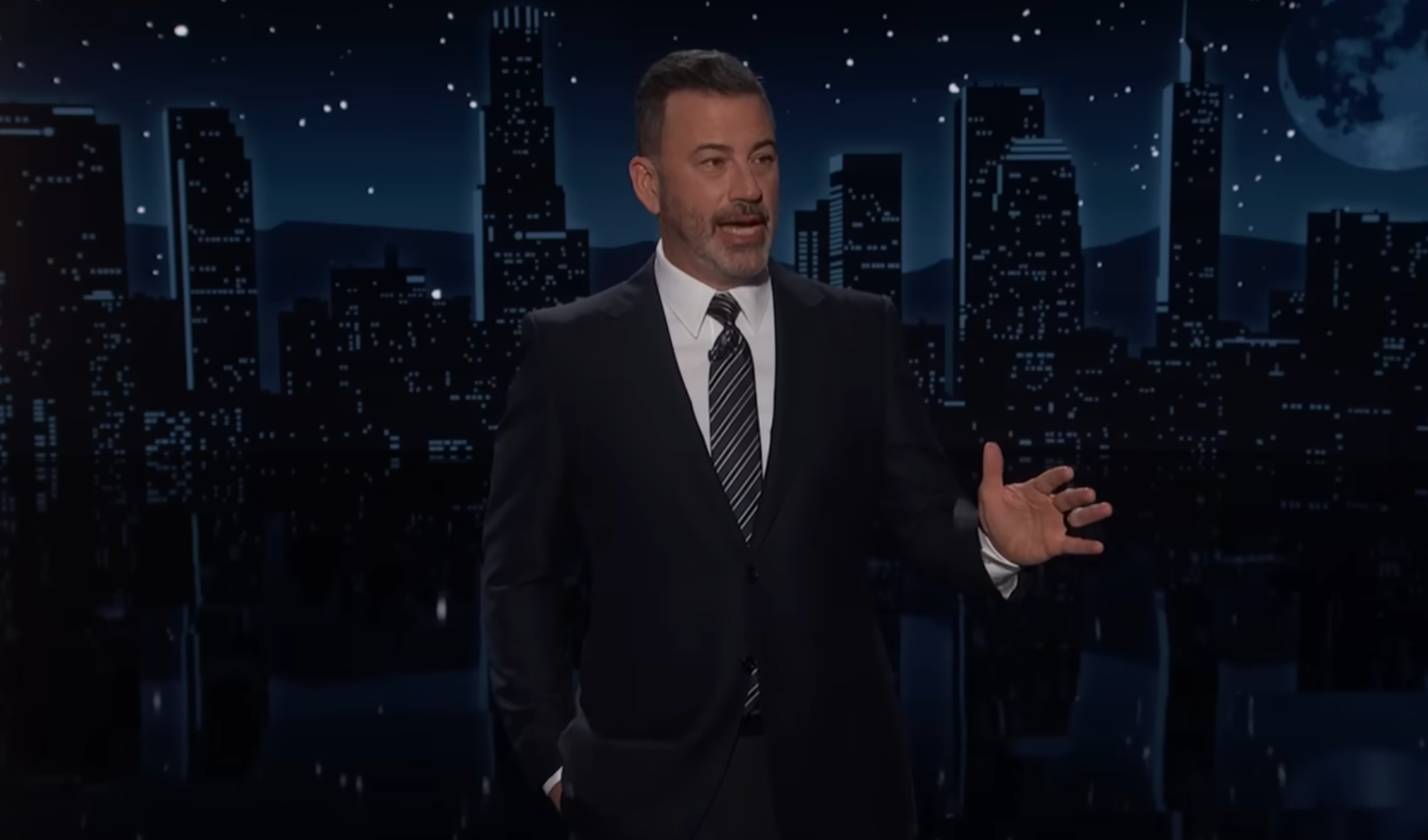 A person in a suit is speaking on a stage with a city skyline backdrop and a starry night sky
