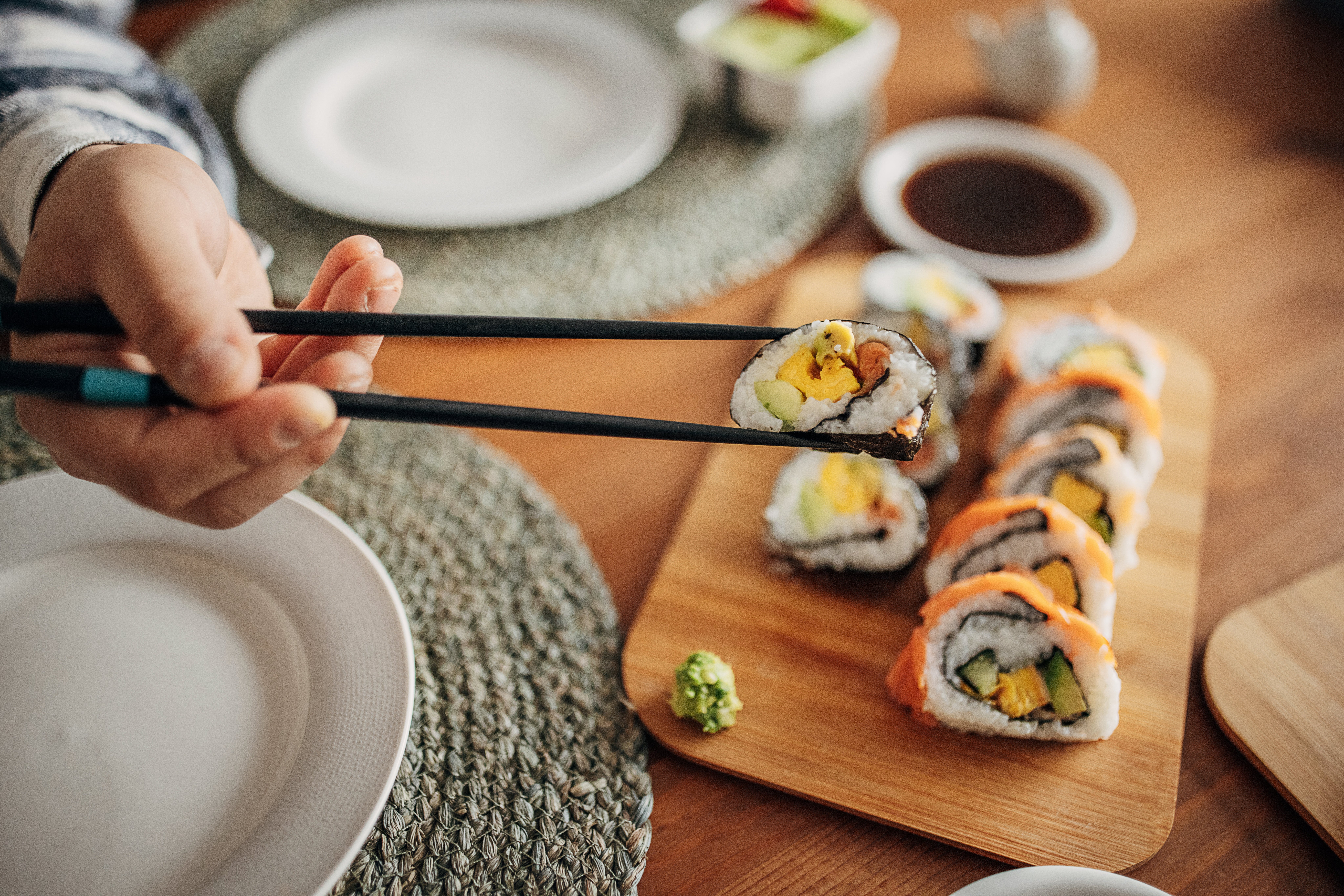 Sebuah tangan menggunakan sumpit untuk memegang gulungan sushi di atas papan kayu dengan berbagai potongan sushi, wasabi, dan saus di dekatnya