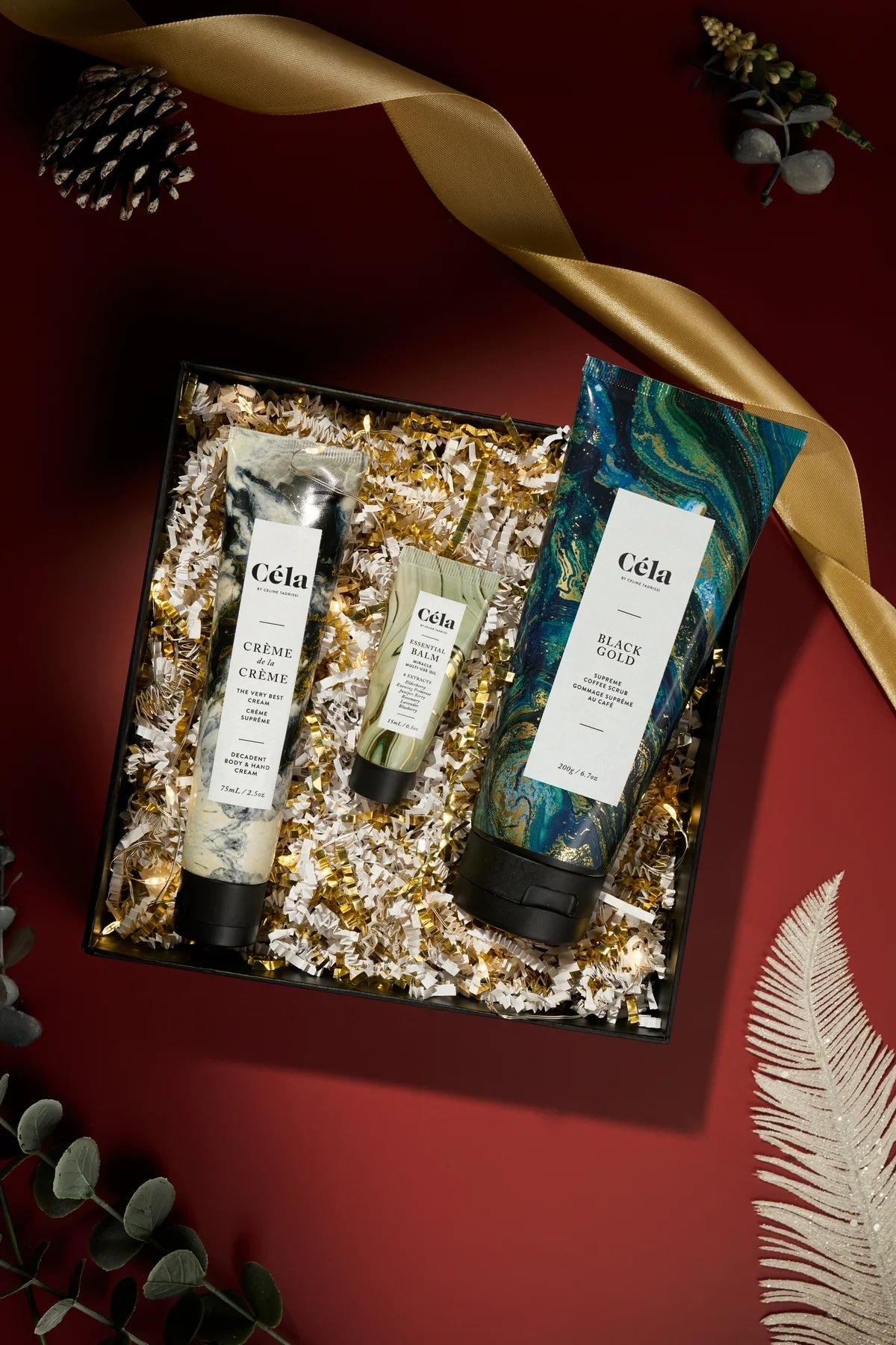 Gift box with three Cela skincare products surrounded by decorative foliage and a gold ribbon, displayed on a festive background