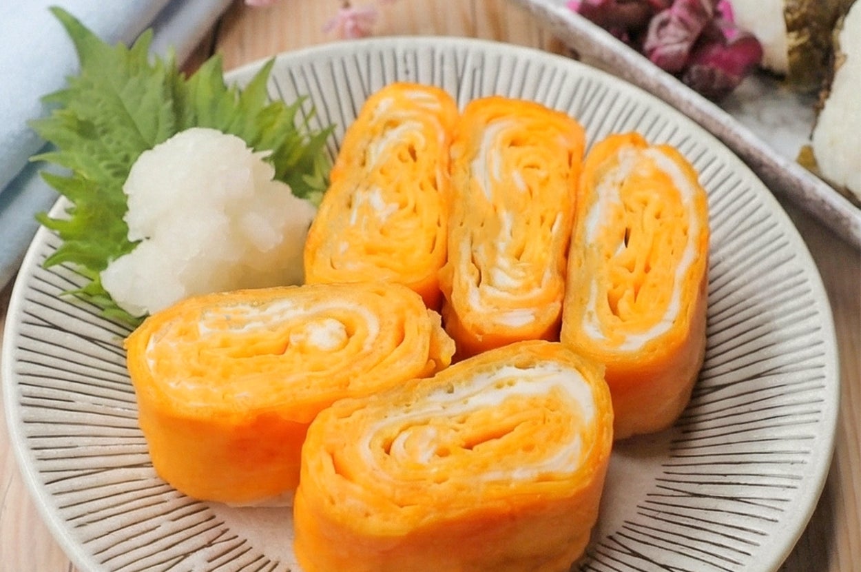 卵焼きが5切れ、葉と大根おろしと共に皿に盛られている。背景におにぎり。