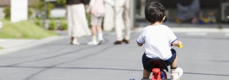 子供が三輪車に乗って住宅街を走っている。家族が道を歩いている。