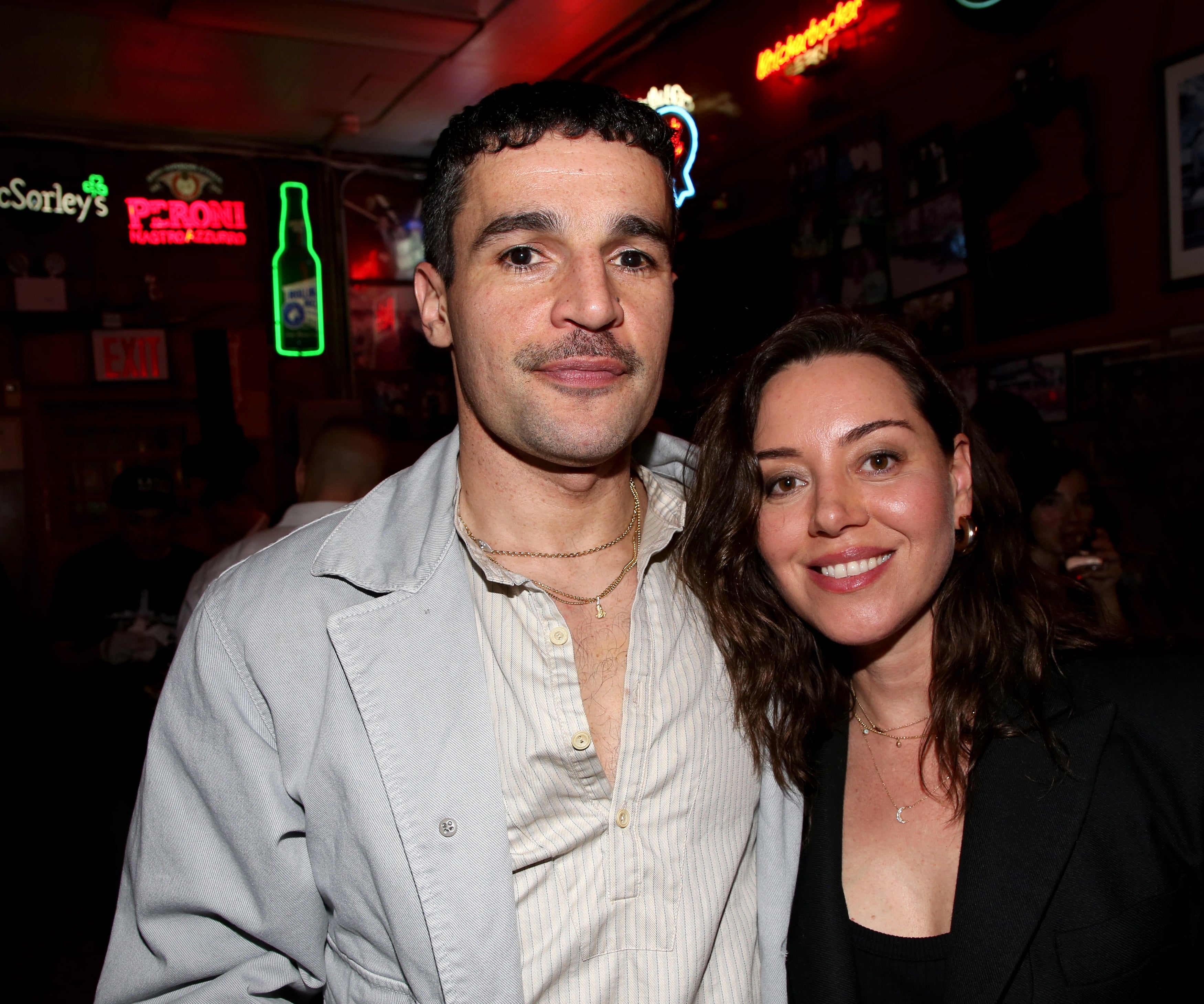 Two people stand together, smiling in a dimly lit venue. One wears a light jacket and shirt, the other a dark outfit, both with casual hairstyles