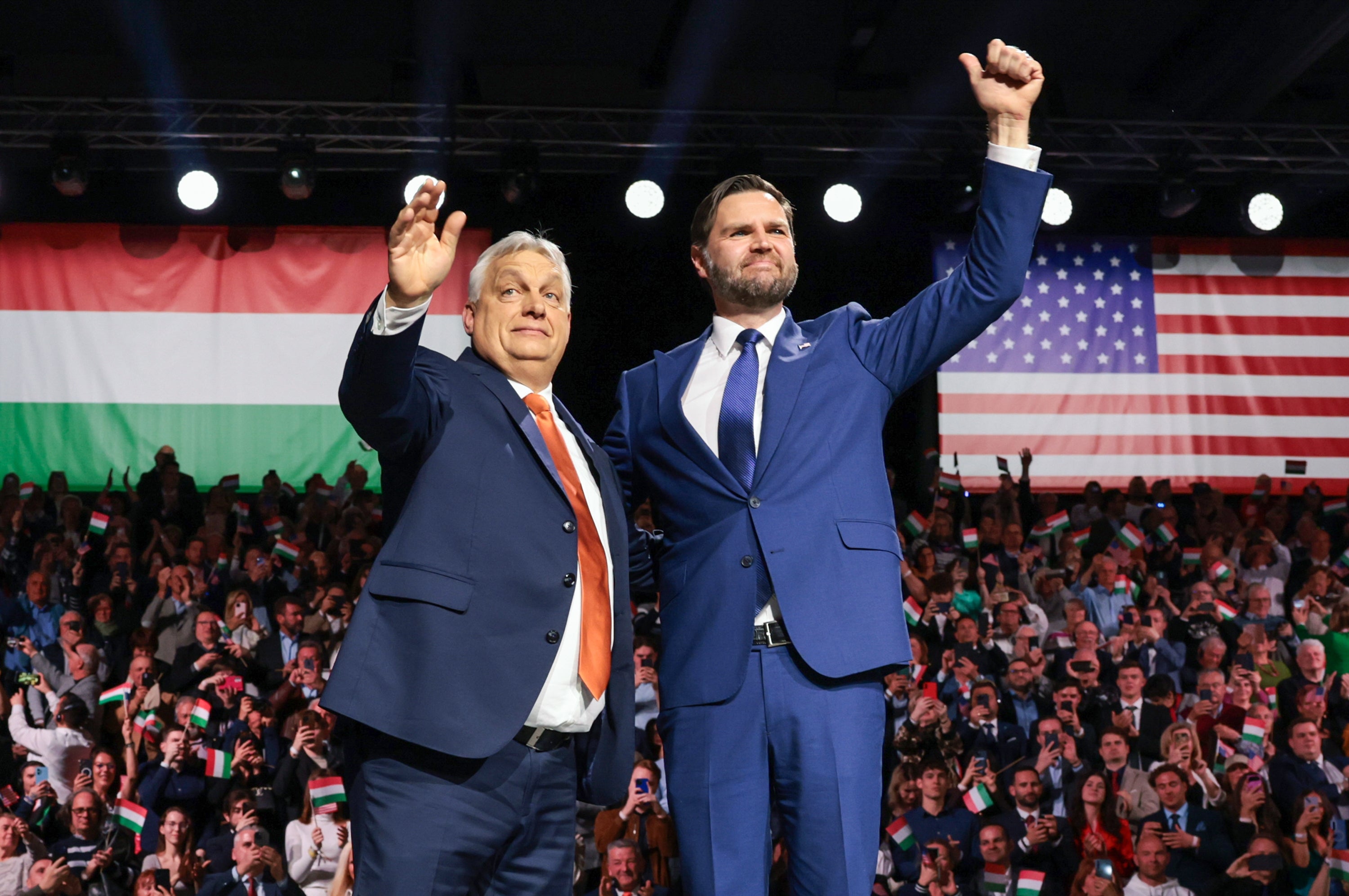 Two men successful suits lasting connected stage, waving to a crowd, with Hungarian and U.S. flags successful the background