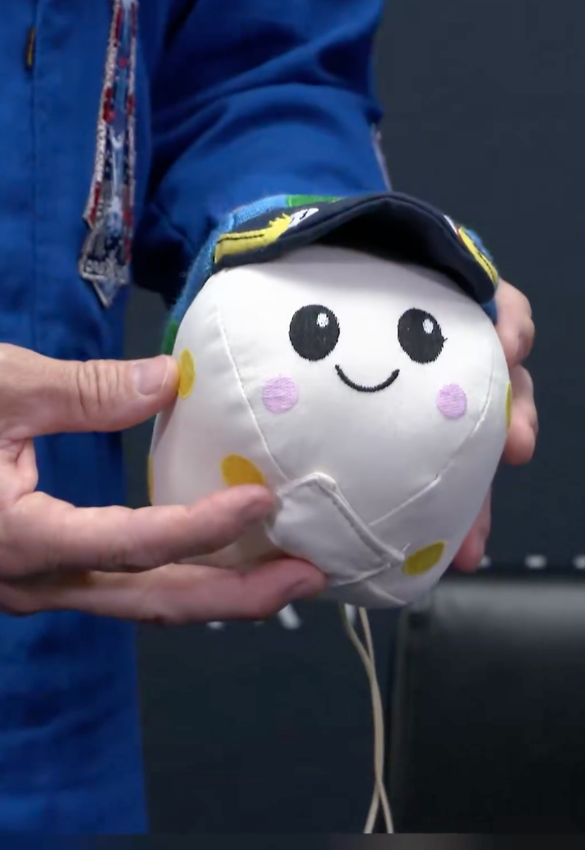Astronaut holds a plush, smiling artifact with a cap, featuring elemental facial details and patterns