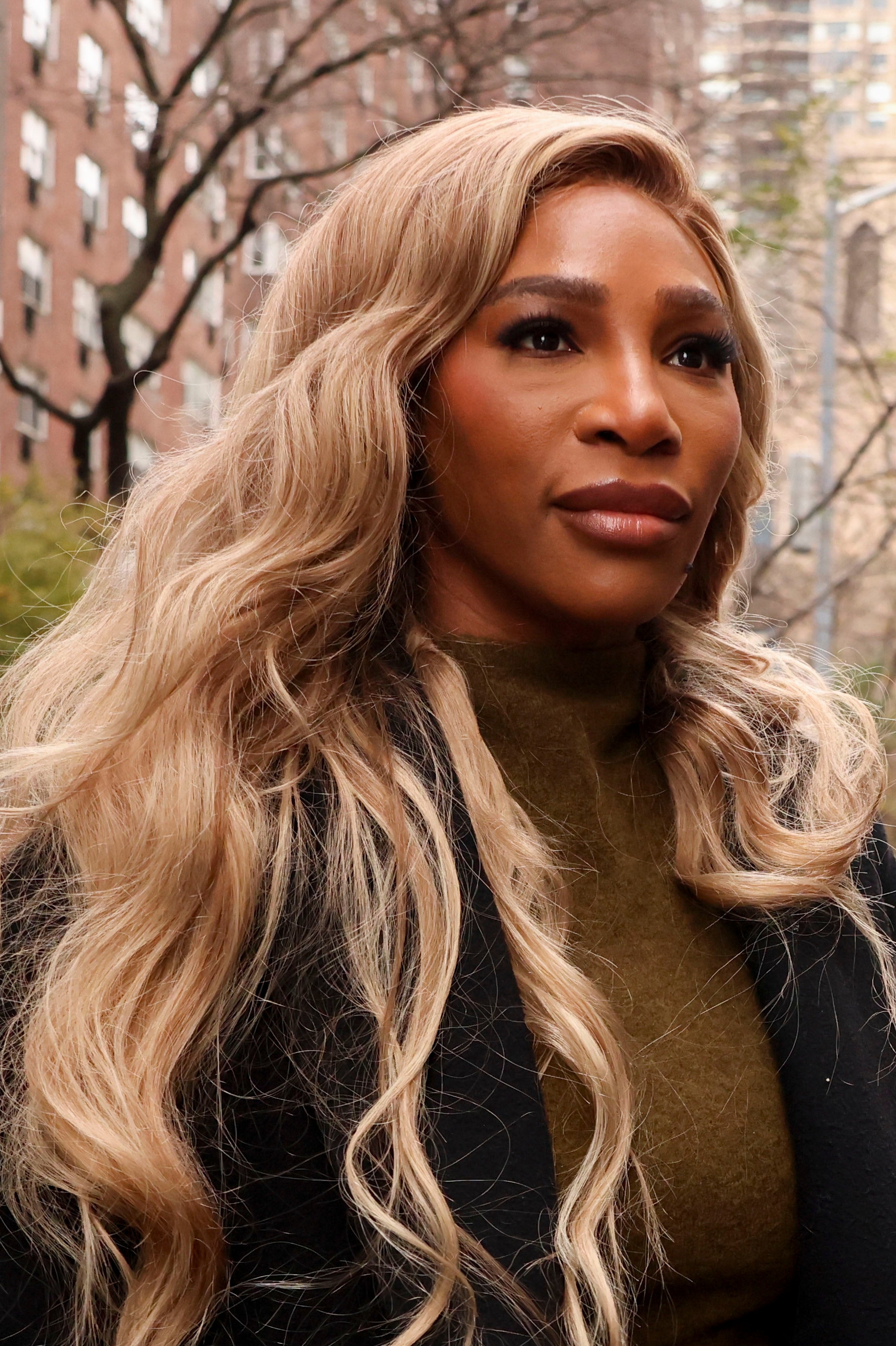 Person with agelong  wavy hairsbreadth  stands outdoors, wearing a acheronian  overgarment  implicit    a high-neck top. Urban inheritance  with trees and buildings visible